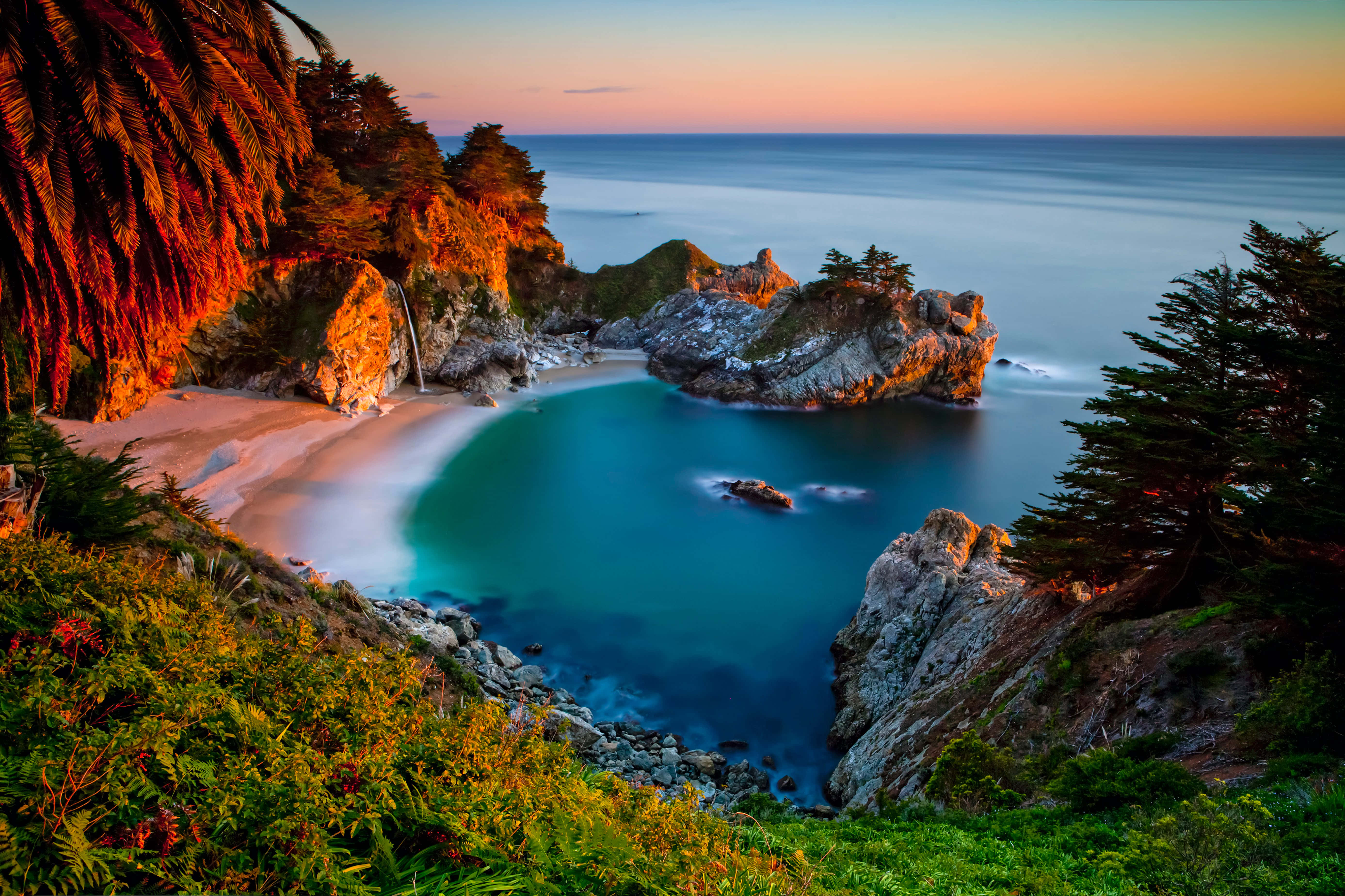 Big Sur 桌布 壁紙 — 日落時分的寧靜海灣，碧綠的海水繞著岩石峭壁流淌，茂密的綠植在溫暖的光線中閃耀。