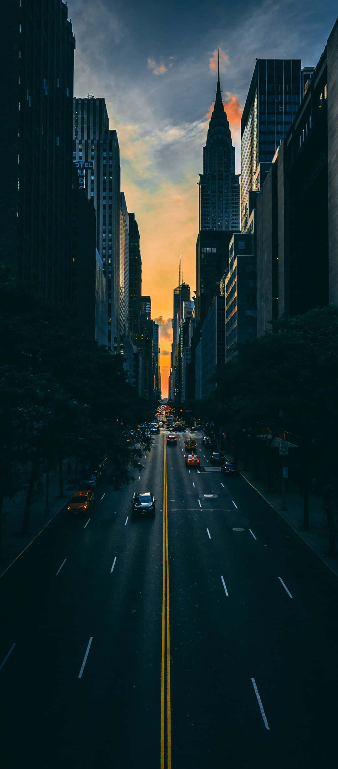 1080x2640 桌布 壁紙 — 日落時分的戲劇性城市峽谷，高聳的摩天大樓框住繁忙街道上方發光的天空。