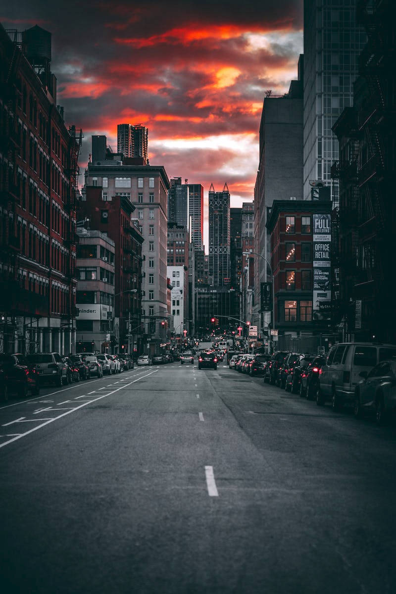 City Street 桌布 壁紙 — 日落時分的戲劇性城市街道，火橙色雲層高聳於高樓大廈和停泊的汽車之上。