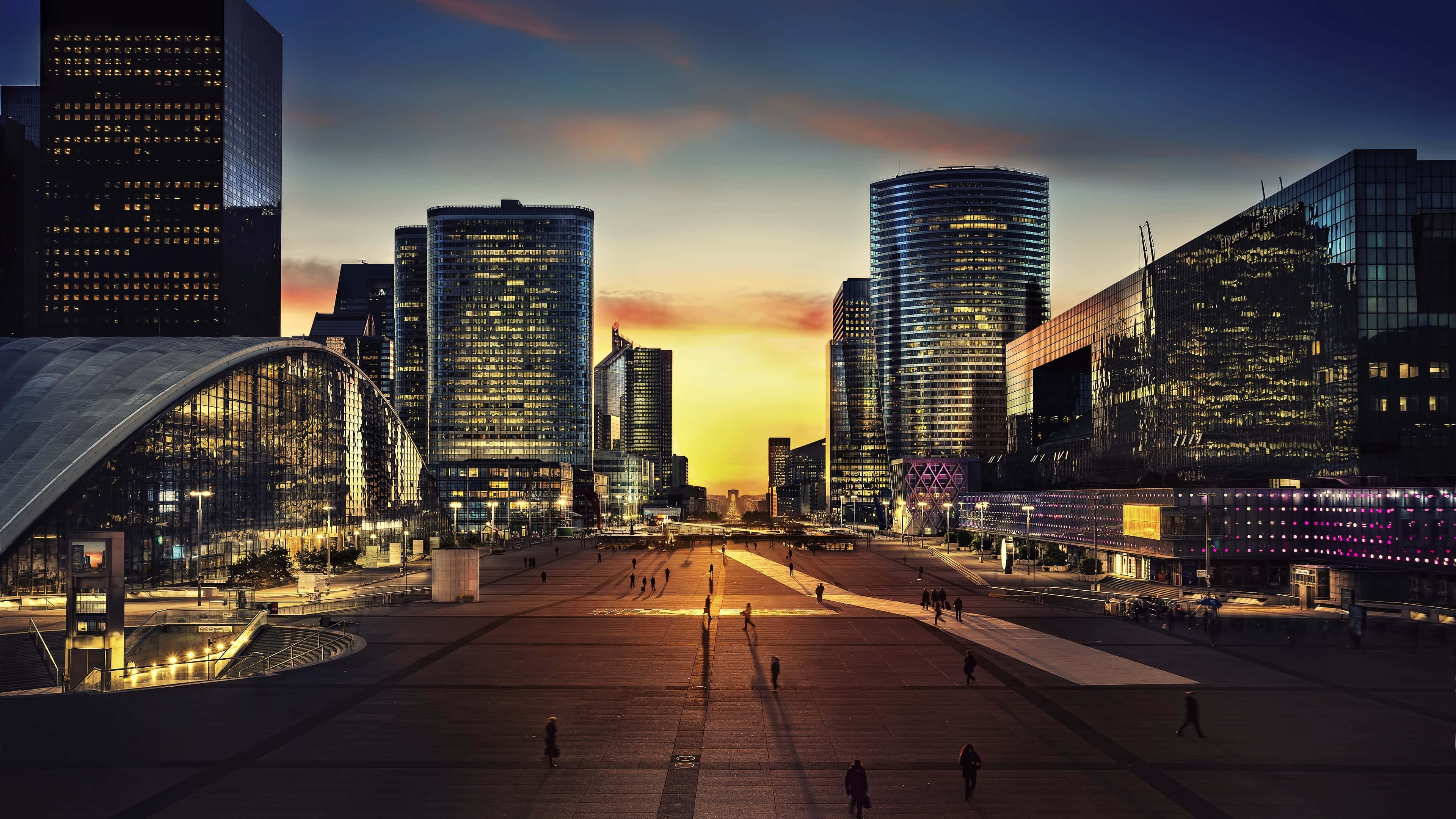Paris At Night 桌布 壁紙 — 日落時分的未來城市景觀，高聳的玻璃摩天大樓和擠滿行人的寬闊廣場。