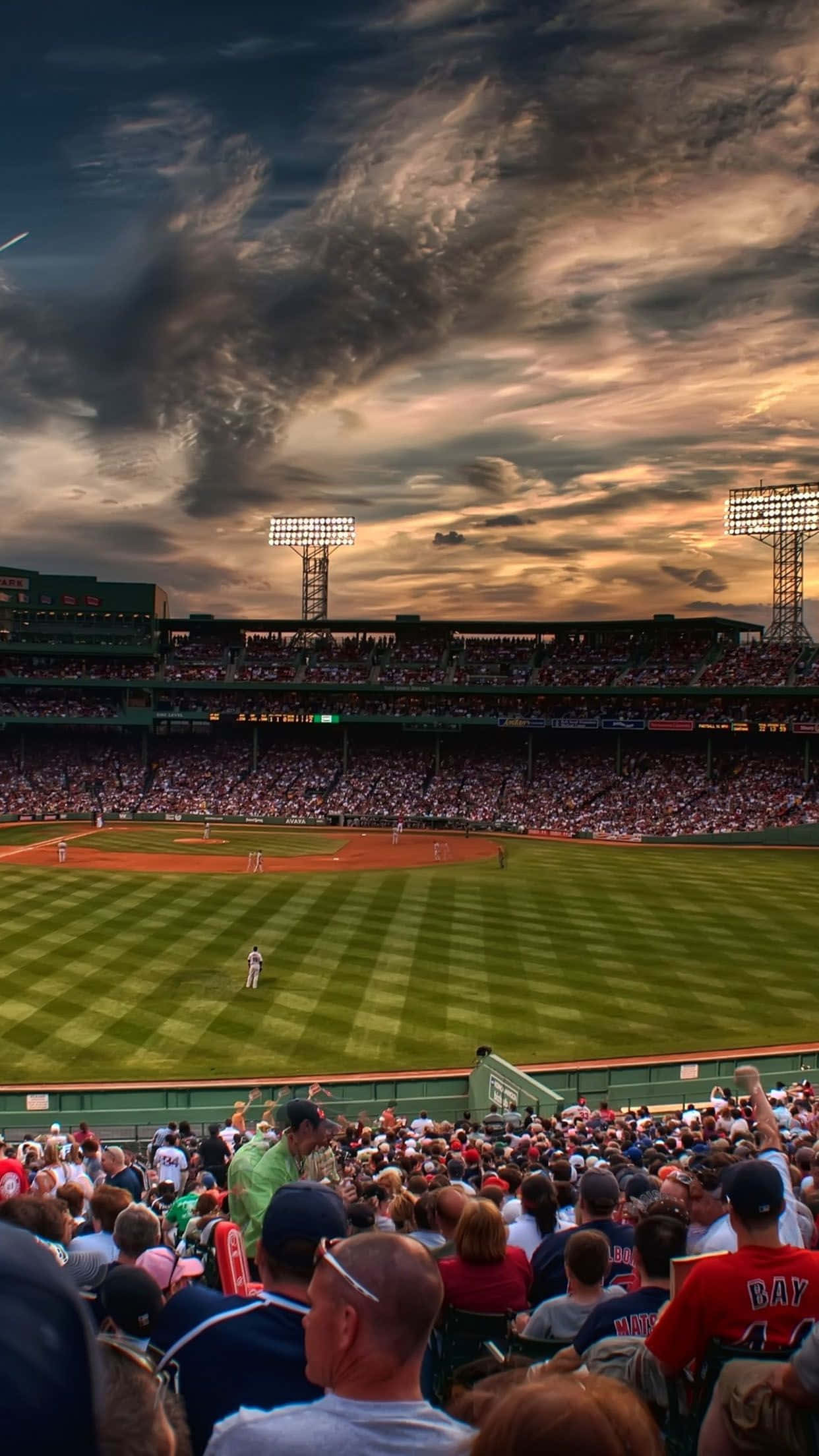 Baseball Stadium 桌布 壁紙 — 日落時分的棒球場，人群在戲劇性的雲層下觀看球員在修剪整齊的場地上比賽。