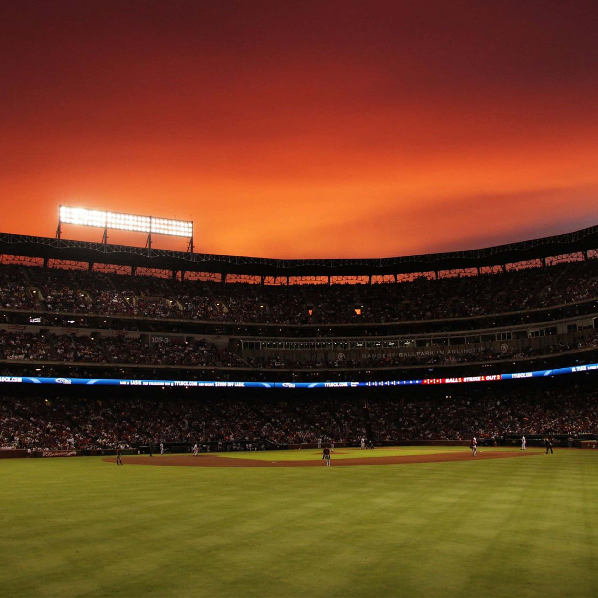 Baseball Field 桌布 壁紙 — 日落時分的棒球場，火紅的橙色天空和發光的燈光照亮球場和擠滿觀眾的看台。