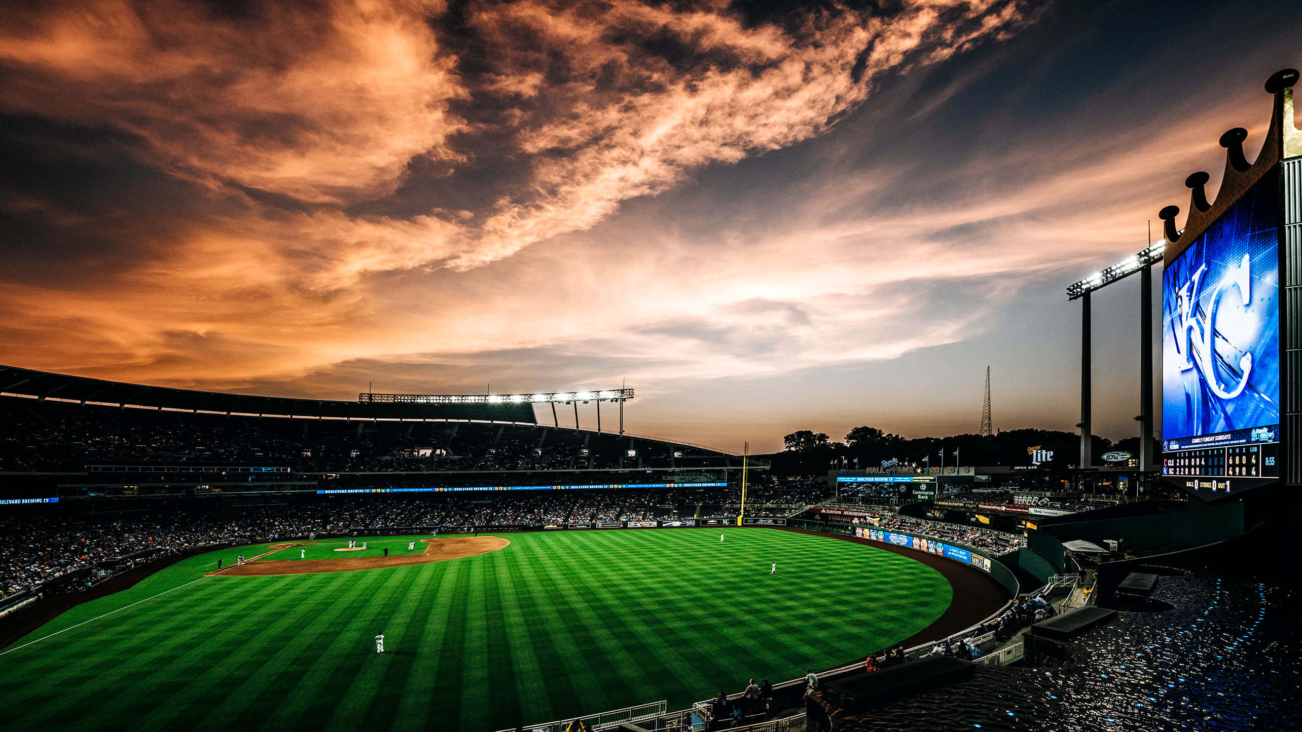 Stadium 桌布 壁紙 — 日落時分的棒球場，綠色球場上方有鮮豔的橙色雲彩和發光的記分牌。
