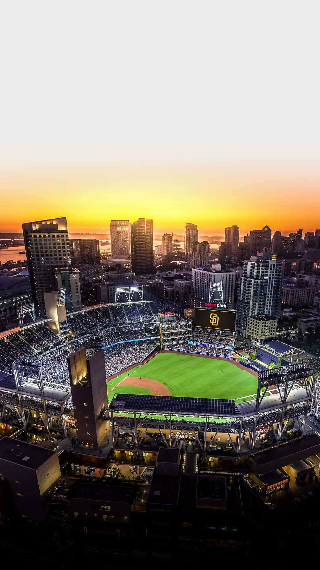 Stadium 桌布 壁紙 — 日落時分的棒球場鳥瞰圖，橙紅色天空發光，城市天際線在背後。