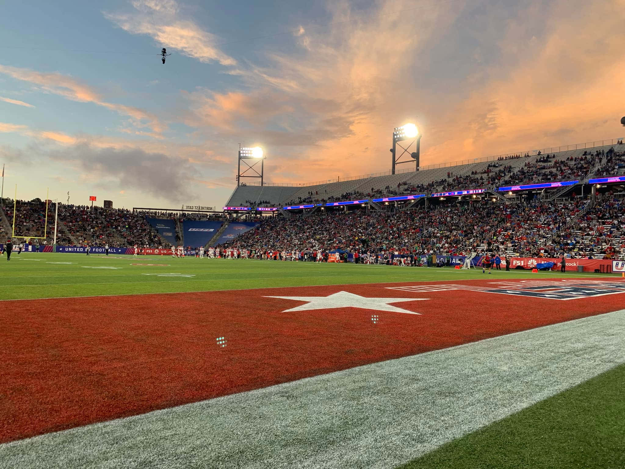 Usfl 桌布 壁紙 — 日落時分的足球場，大量人群在閃爍燈光下歡呼，球員們在場上列隊。
