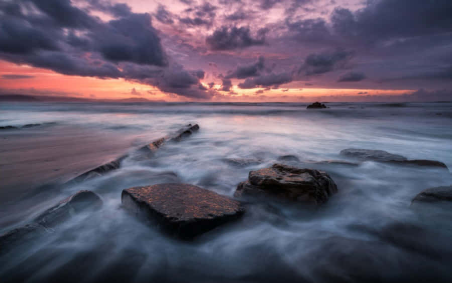 Dark Sky 桌布 壁紙 — 日落時分的長時間曝光海景，平滑的水流在深色岩石周圍流動，天空呈現戲劇性的紫色和橙色。