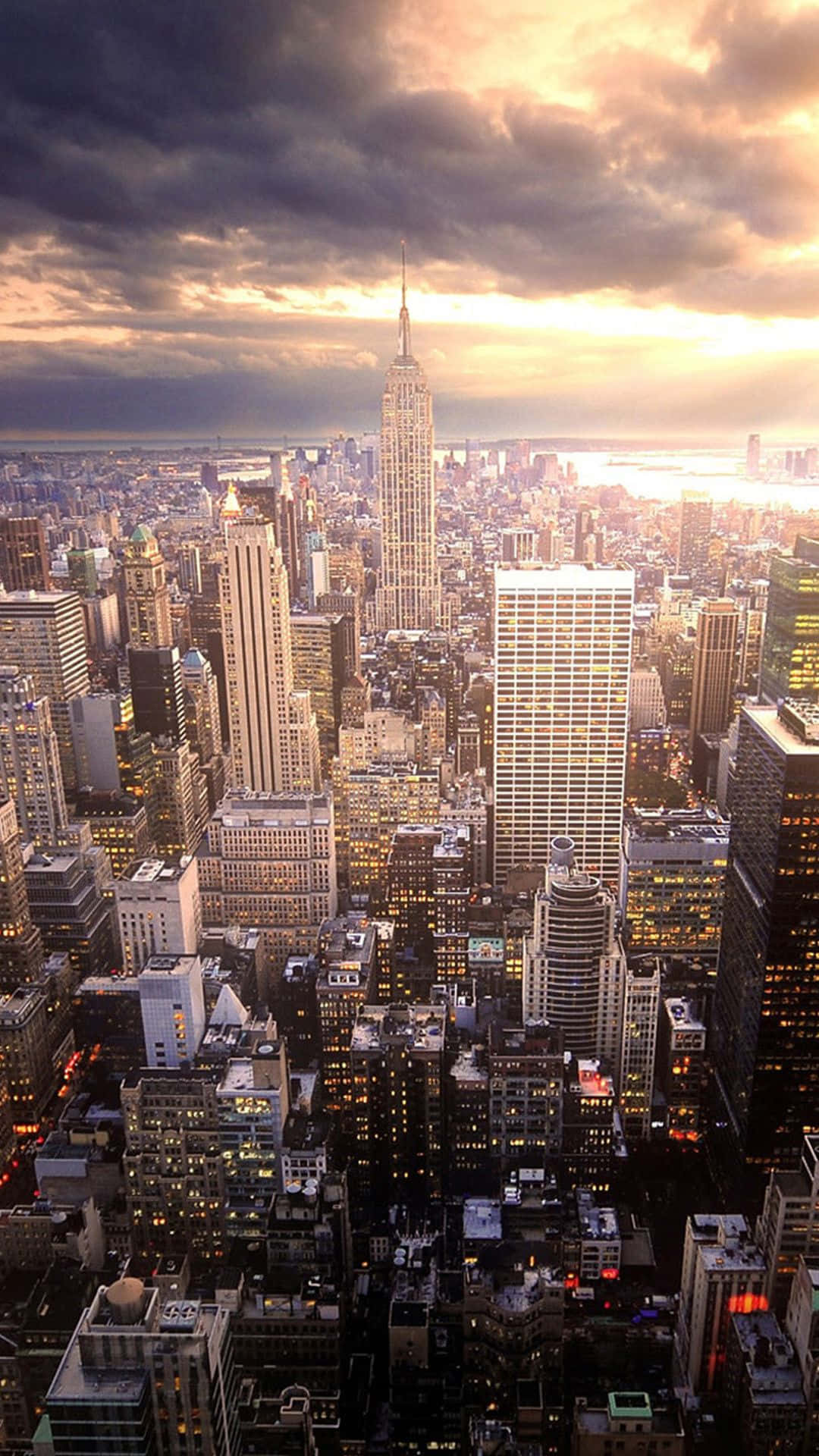 New York City Night iPhone 桌布 壁紙 — 日落時分紐約市天際線的鳥瞰圖，帝國大廈在密集的城市建築上方聳立，天空佈滿戲劇性的雲層。