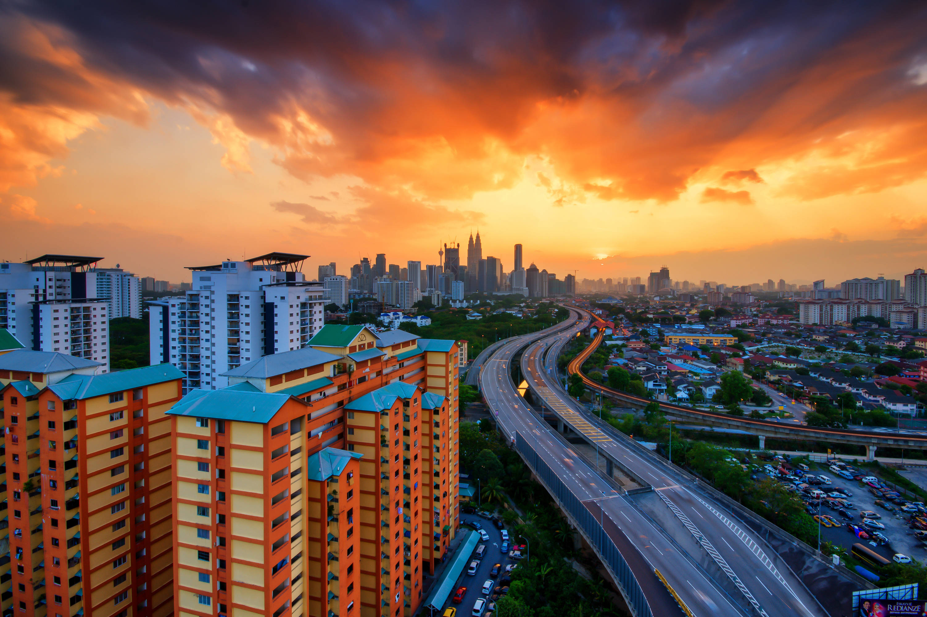 Malaysia 桌布 壁紙 — 日落時分繁忙城市景觀的鳥瞰圖，高聳建築物和繁忙高速公路之上有鮮豔的橙色雲彩。