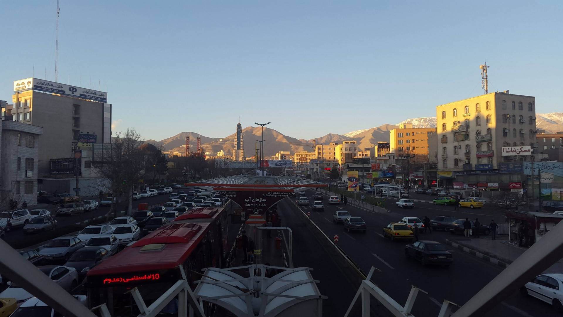 德黑蘭 桌布 壁紙 — 日落時分繁華的城市街道，背景是白雪覆蓋的山脈，交通在晴朗的藍天下流動。
