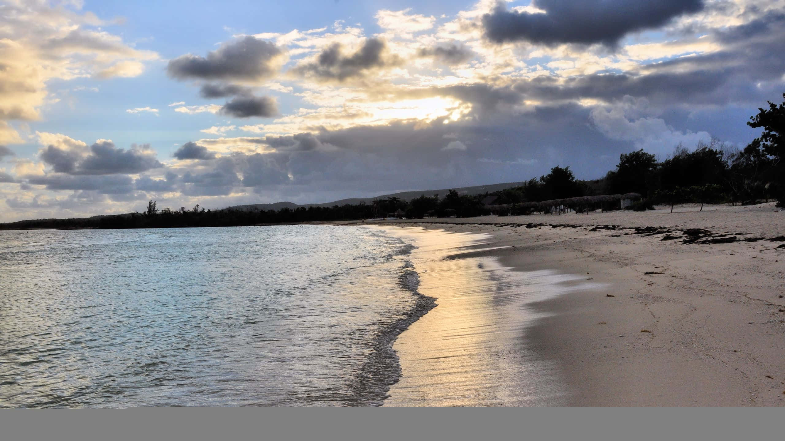 1440p Beach 桌布 壁紙 — 日落用金色雲彩繪畫天空，映照在寧靜的海灘上，溫柔的海浪親吻著白色沙灘。