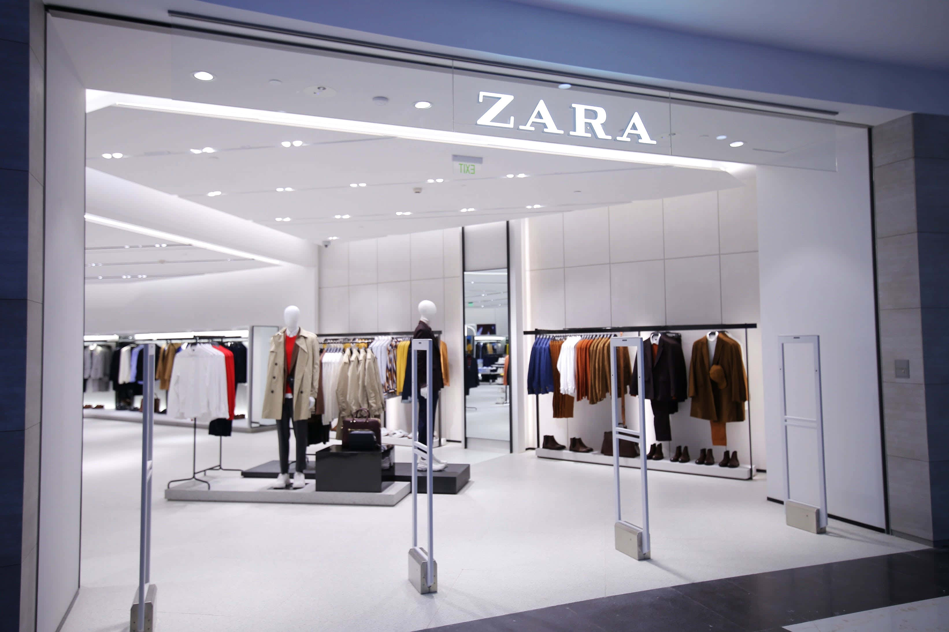 Zara 桌布 壁紙 — 明亮現代的Zara商店入口，展示整齊排列的衣架和模特兒，在潔淨的白色燈光下。
