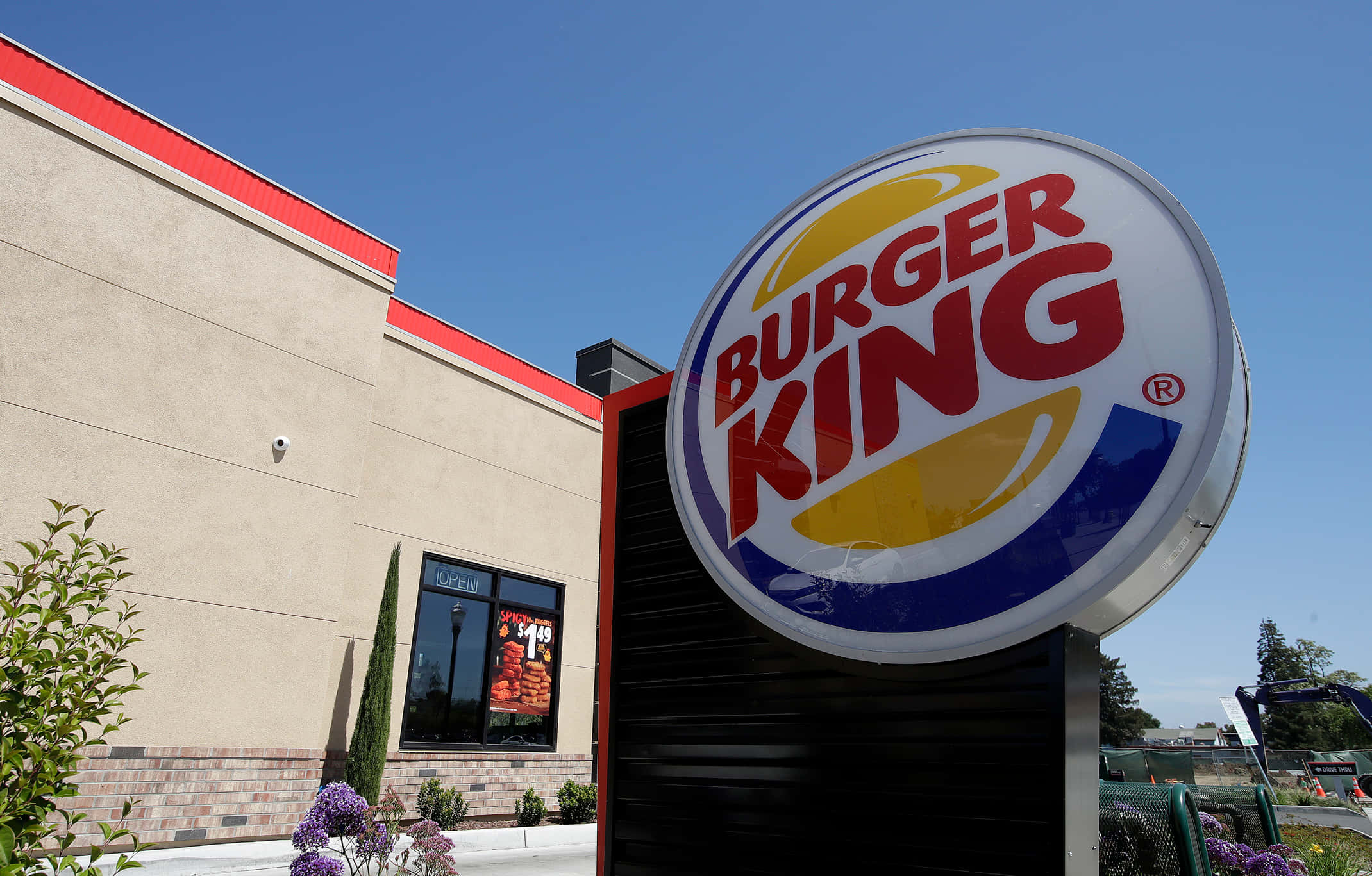 Burger King 桌布 壁紙 — 明亮的Burger King招牌在晴朗的藍天下高高聳立，旁邊是米色餐廳建築，帶有紅色飾邊和附近的綠植。