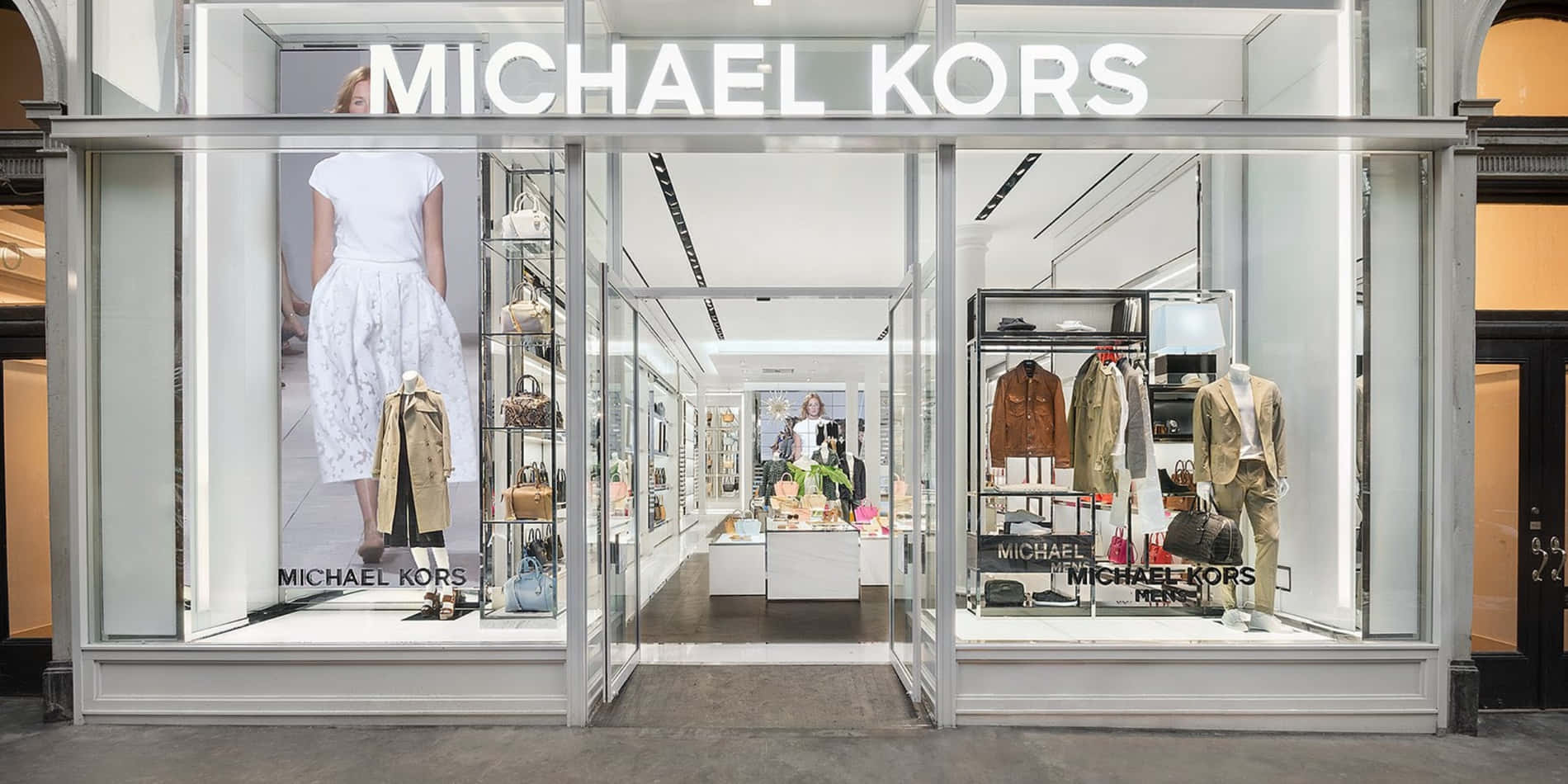 麥可·寇斯 桌布 壁紙 — 明亮的Michael Kors店鋪正面，玻璃窗展示時尚人體模型和商品，現代燈光下。