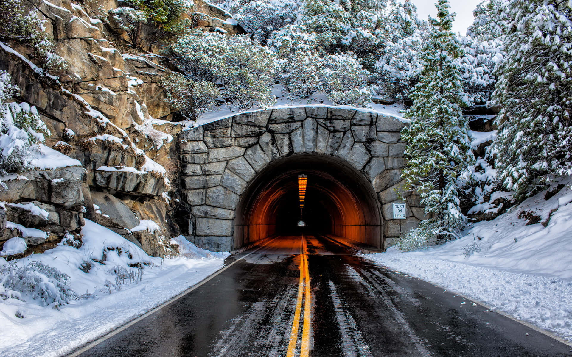 Road 桌布 壁紙 — 明亮的冬日天空下，常綠樹和岩石峭壁環繞著一個積雪的石隧道入口。