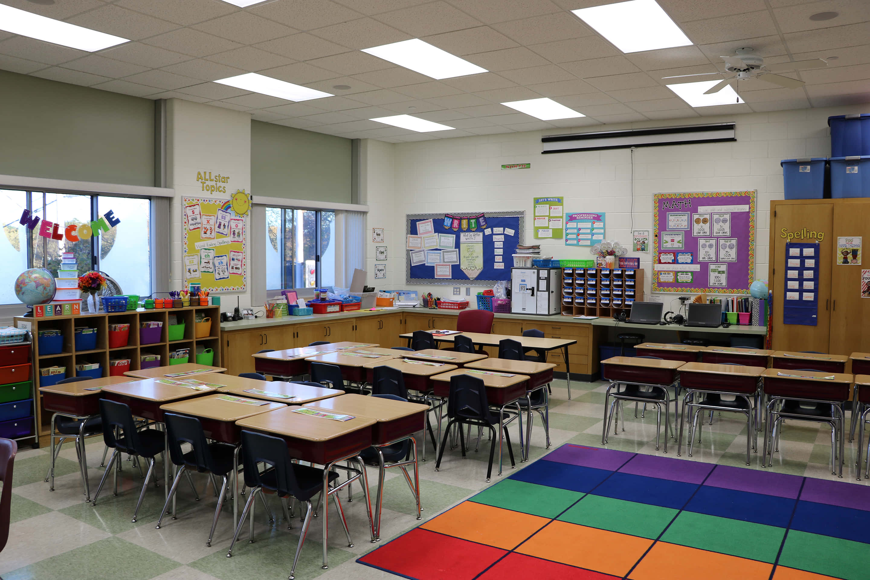 Classroom 桌布 壁紙 — 明亮的初級教室，配有彩色彩虹地毯、書桌、椅子和教育海報，在螢光燈下。