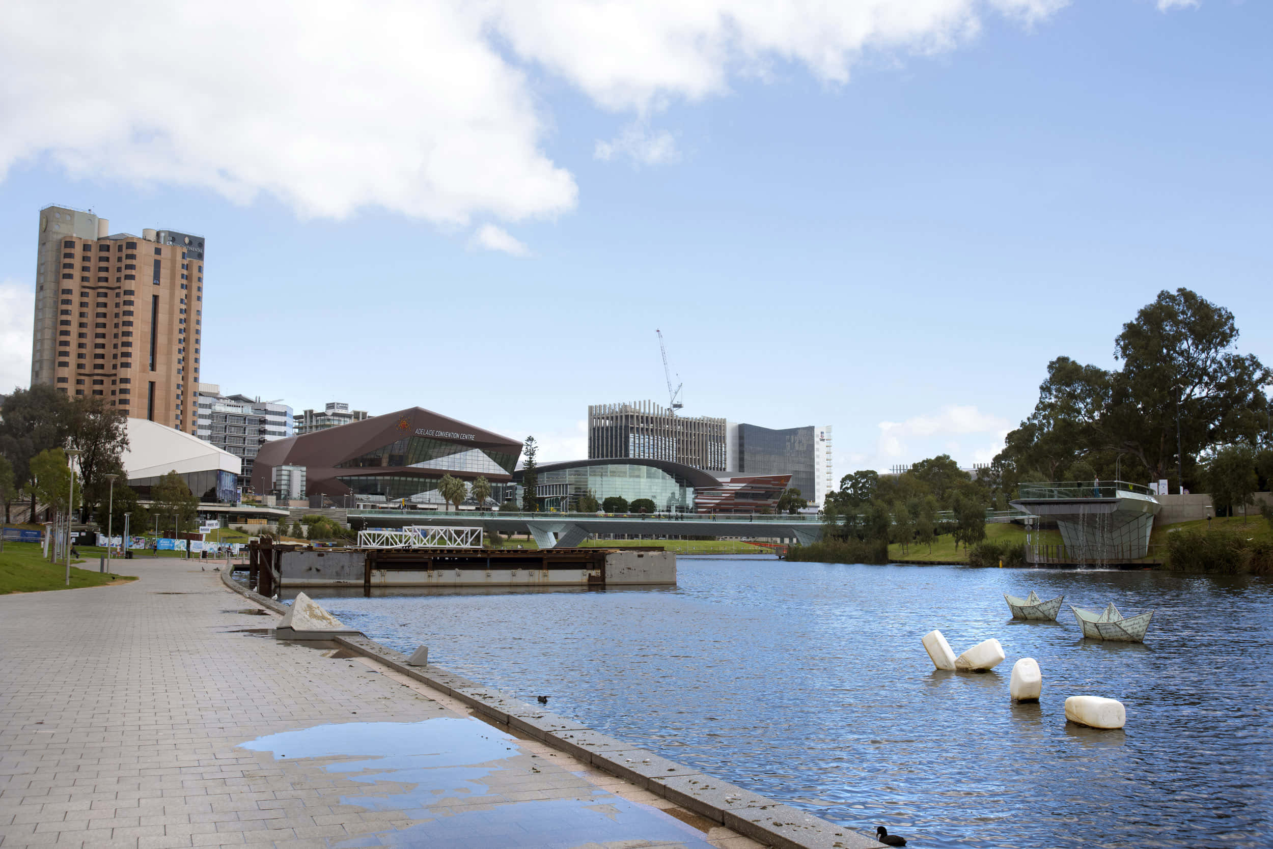 Adelaide 桌布 壁紙 — 明亮的城市海濱場景，有現代建築、橋樑和白色雕塑漂浮在平靜的藍色水面上，天空部分多雲。