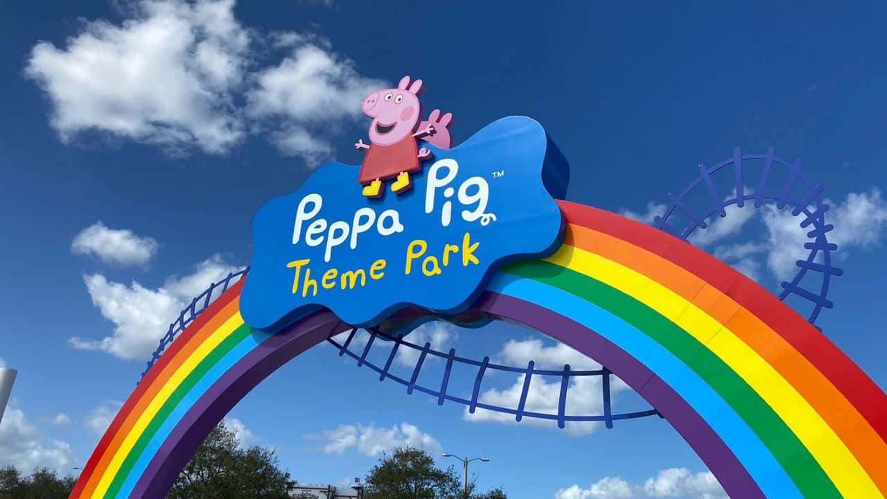 Peppa Pig Family 桌布 壁紙 — 明亮的彩虹拱門，Peppa Pig 在藍色標誌上微笑，Peppa Pig 主題公園，陽光明媚的天空下。