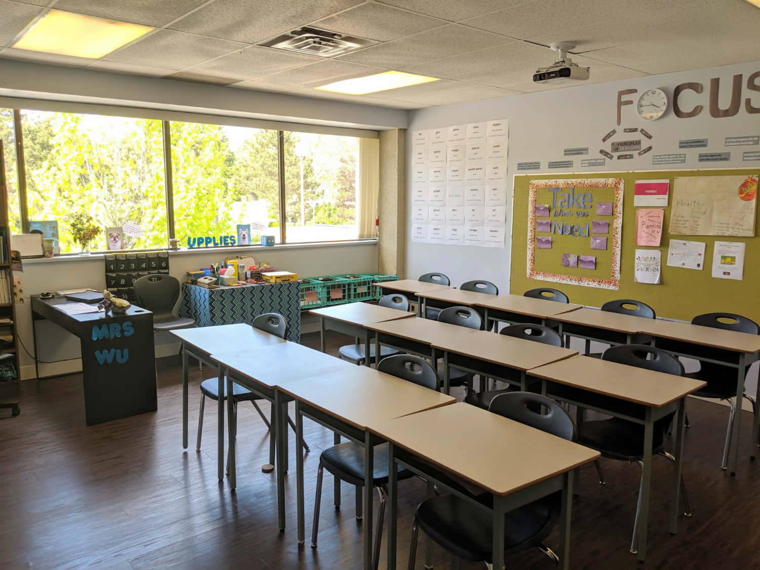 Real Classroom 桌布 壁紙 — 明亮的教室，螢光燈下排列著書桌、椅子和教育海報，窗外可見樹木。