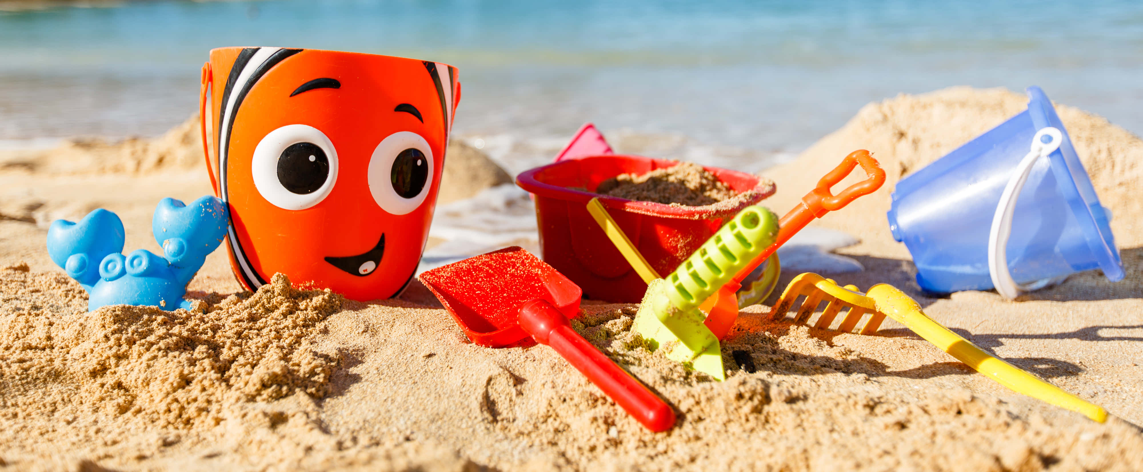 Beach Toys 桌布 壁紙 — 明亮的沙灘玩具，包括Nemo水桶、鏟子、耙子和沙模，放在金色沙灘上，靠近海浪。