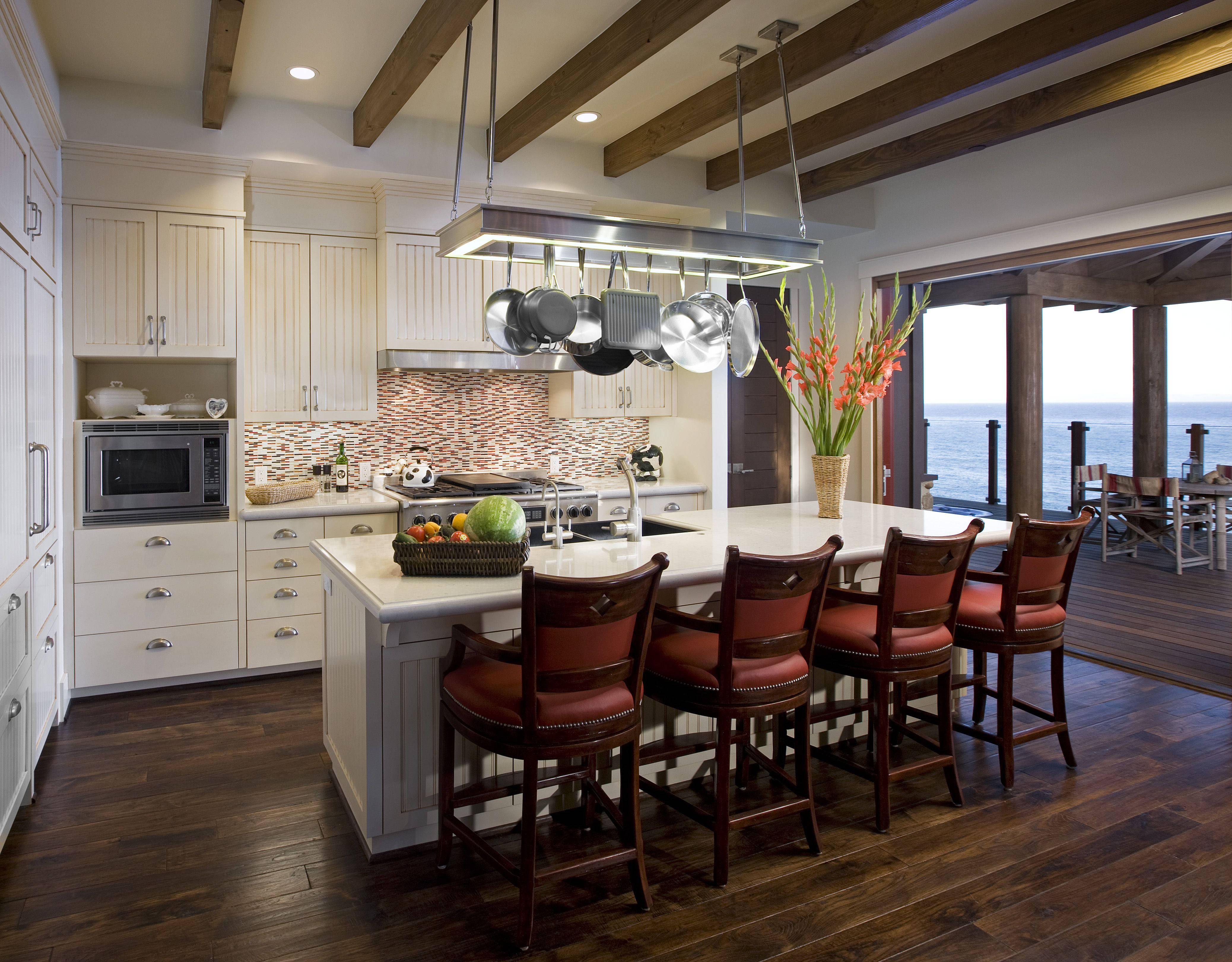 Kitchen Design 桌布 壁紙 — 明亮的沿海廚房，配有白色櫥櫃、深色木地板和紅色皮革吧檯椅，透過大型玻璃門俯瞰海洋。