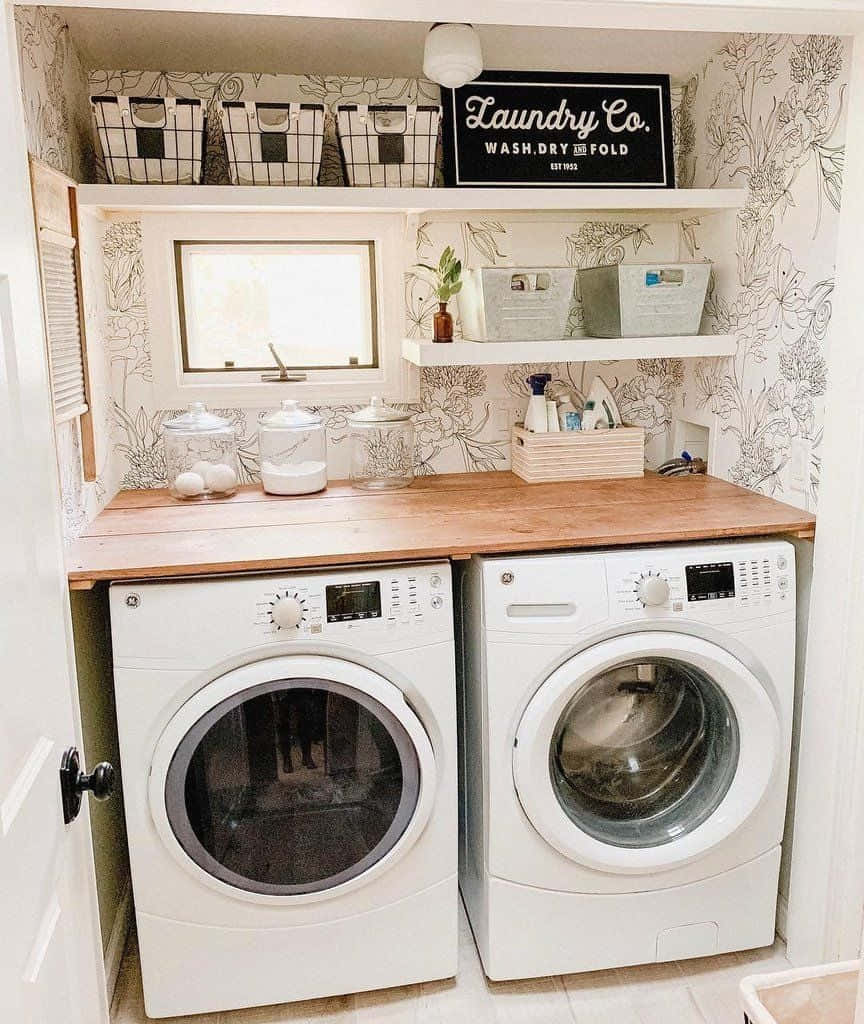 Laundry Room 桌布 壁紙 — 明亮的洗衣房，白色前置式洗衣機位於木製檯面下，花卉壁紙層架上放有籃子和「Laundry Co.」標誌。