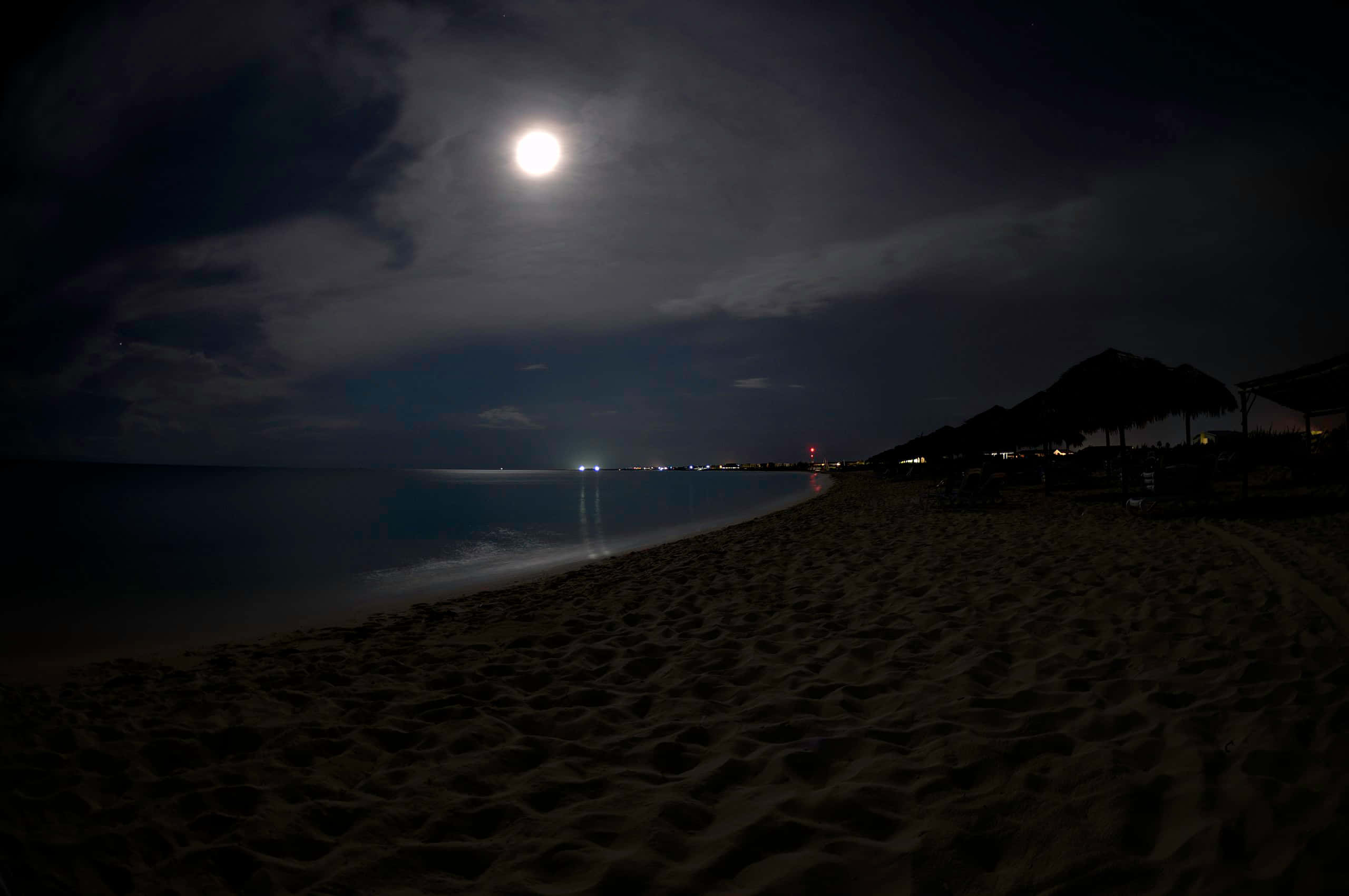海灘 桌布 壁紙 — 明亮的滿月在夜晚將銀色光芒灑在寧靜的海灘上，沙灘呈深色，地平線上有遠處的燈光。