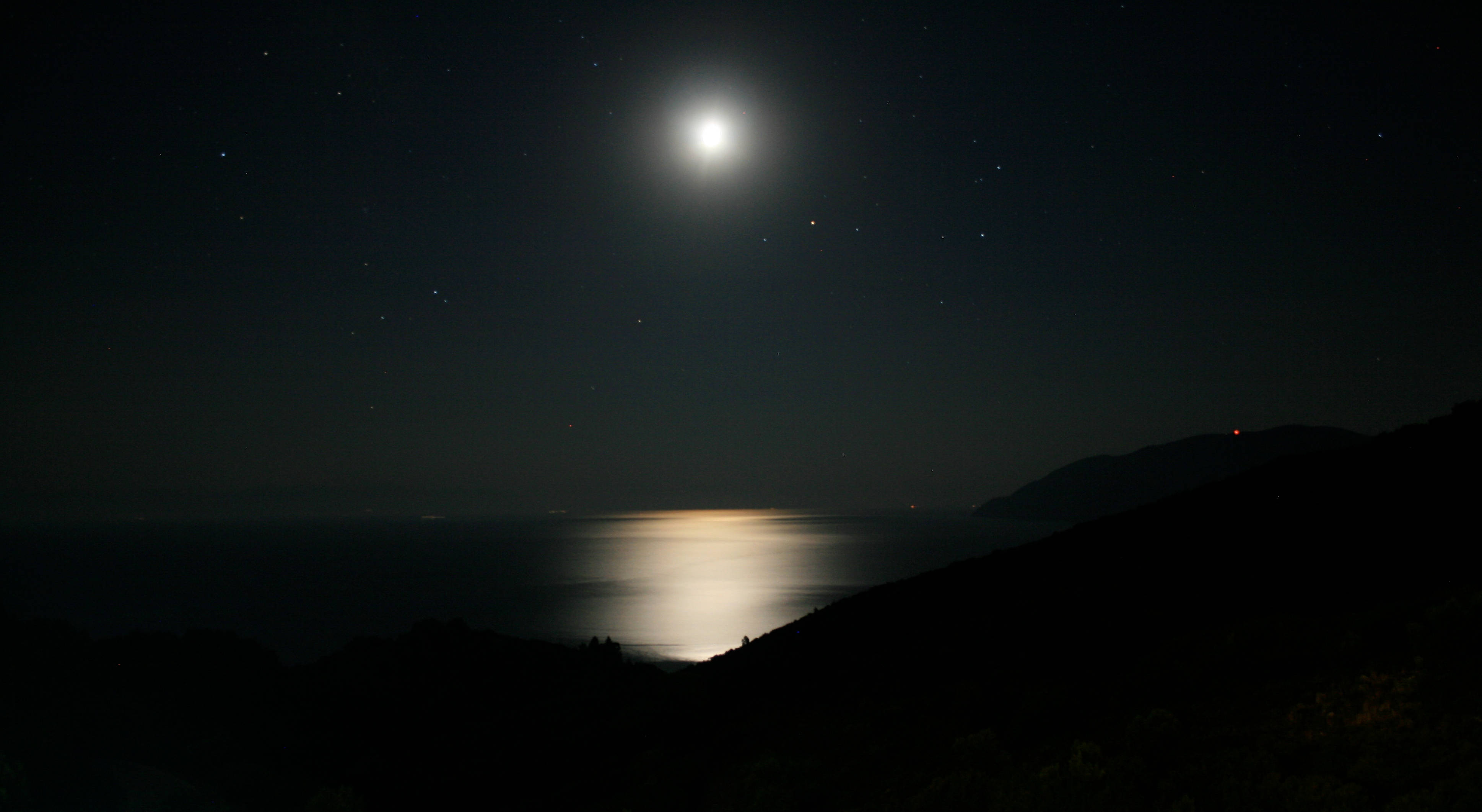 Moonlight 桌布 壁紙 — 明亮的滿月在平靜的海面上投射出一條閃爍的路徑，夜空繁星點點，黑暗的山丘輪廓分明。