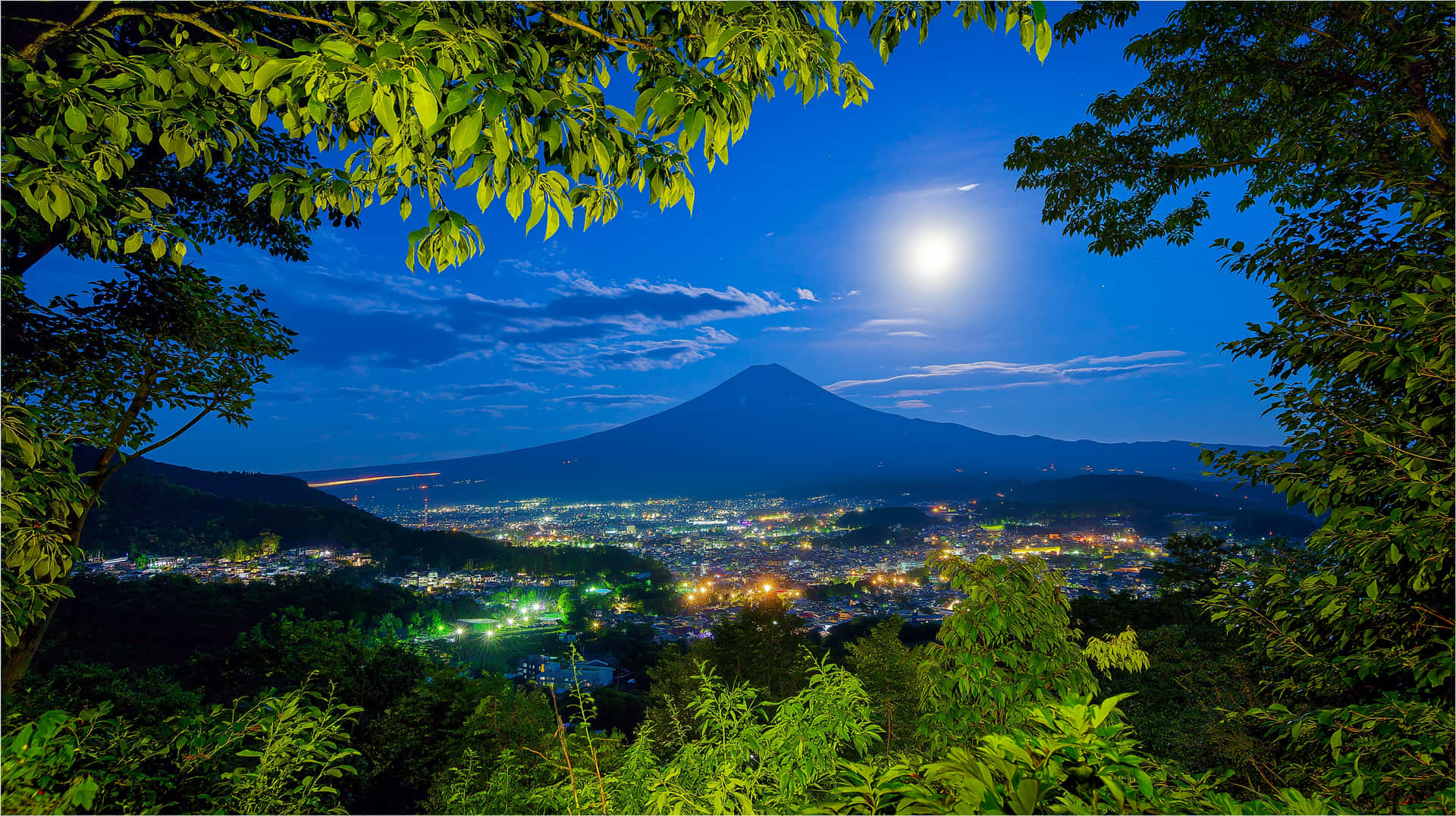 Moonlit Mount Fuji Over Glowing City at Night 壁紙 — 明亮的滿月照耀著富士山，下方城市燈光閃爍，被鮮綠的樹葉環繞。