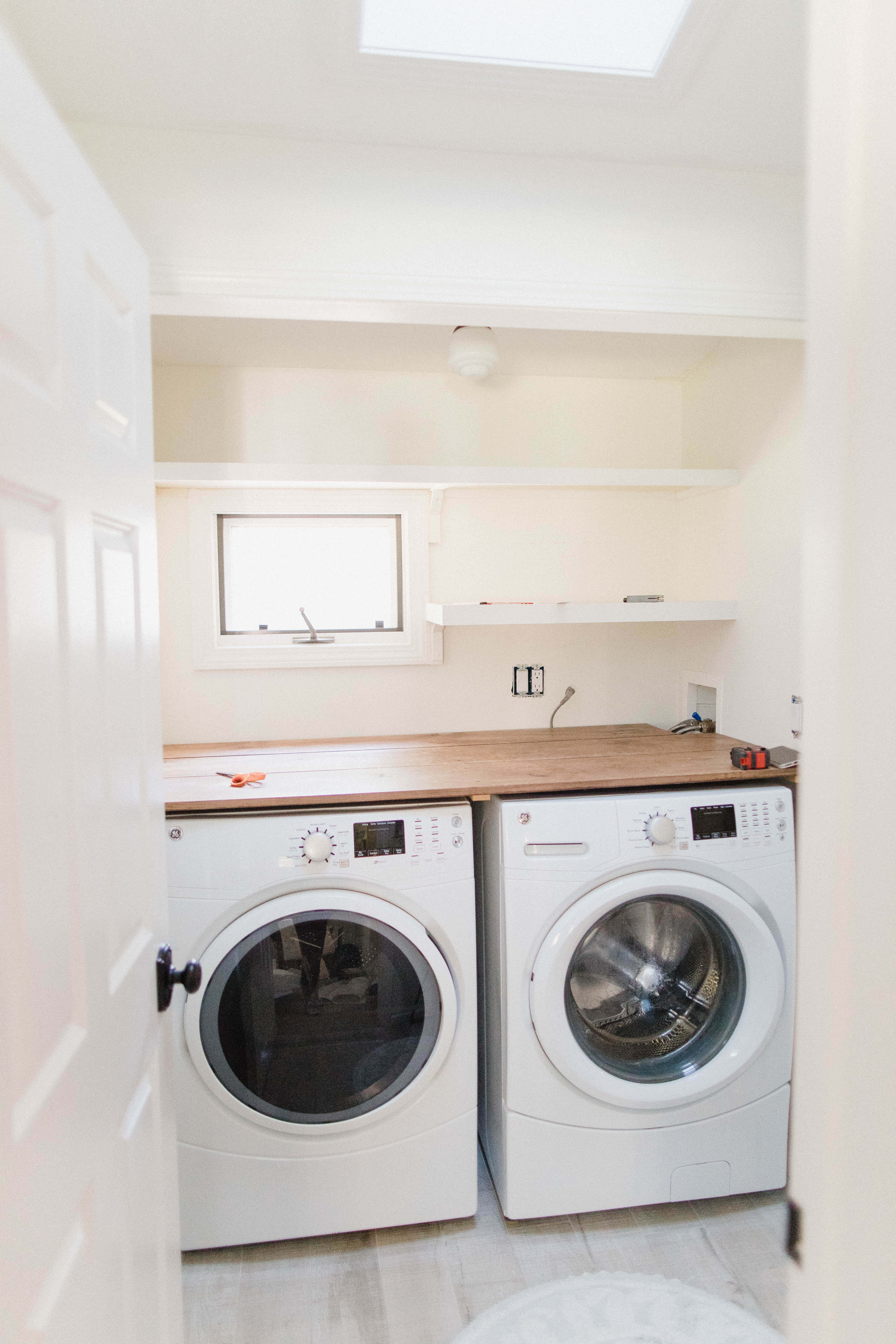 Laundry Room 桌布 壁紙 — 明亮的簡約洗衣房，木製檯面和天窗下設有兩部白色前置式洗衣機。