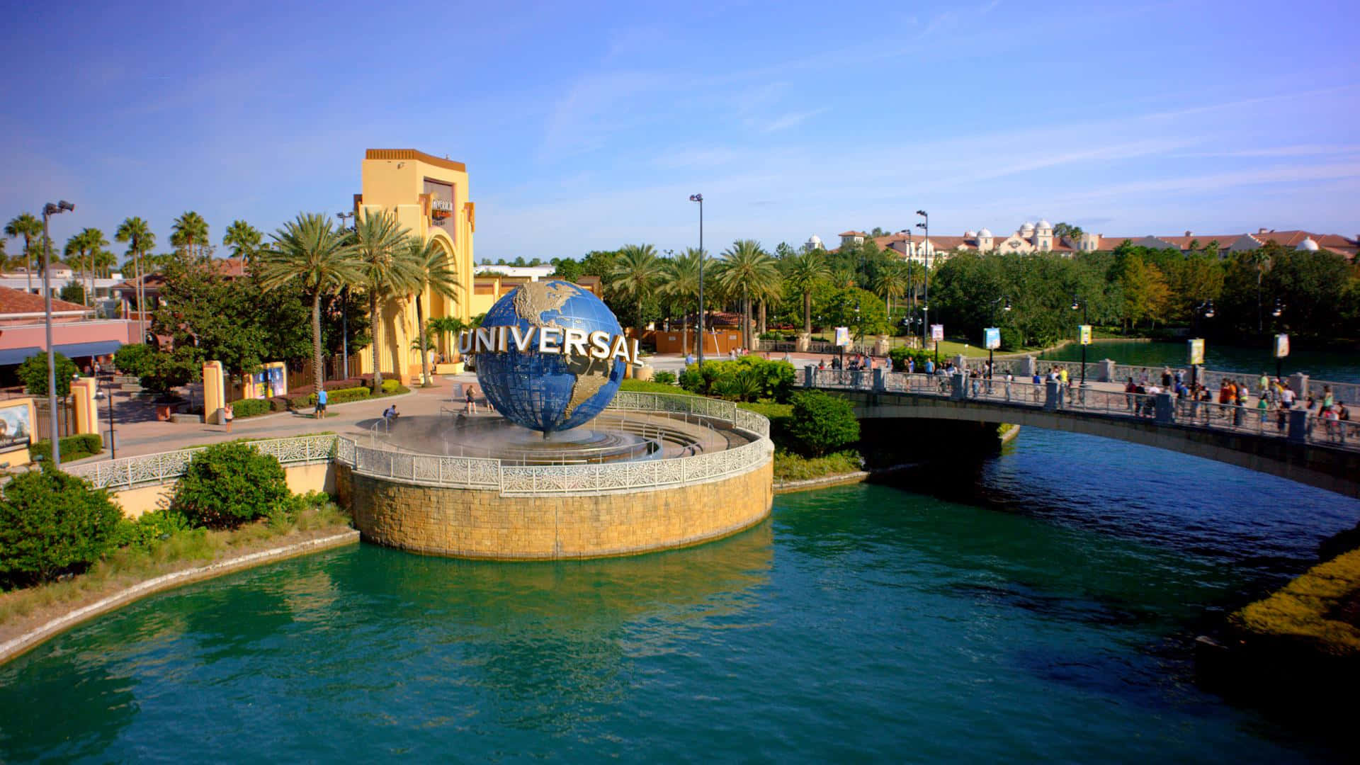 Universal 桌布 壁紙 — 明亮的藍天下，標誌性的環球影城地球標誌被碧綠的海水和棕櫚樹環繞，遊客在橋上漫步。