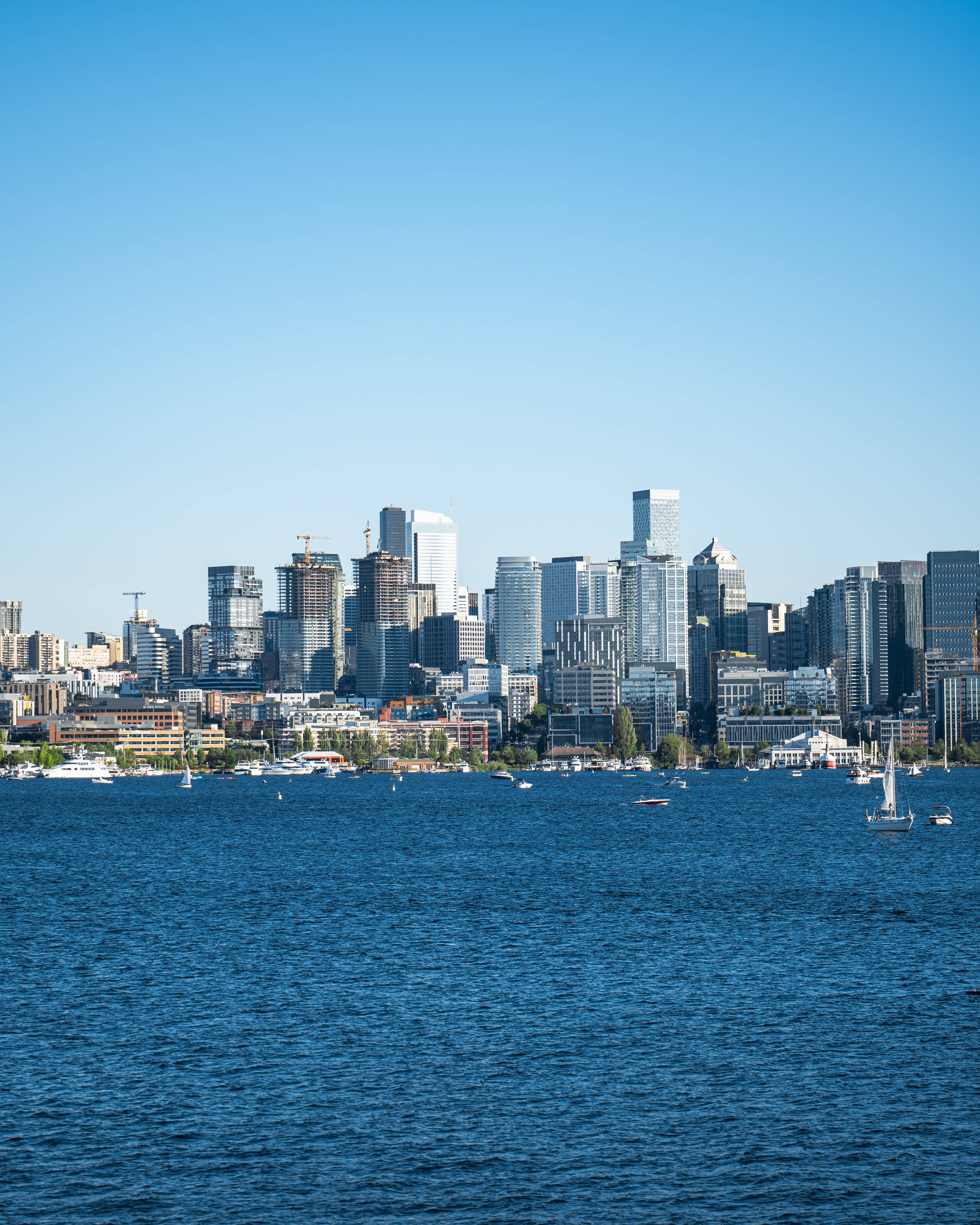 seattle 4K 桌布 壁紙 — 明亮的藍天下，現代城市天際線的玻璃摩天大樓倒映在點綴著帆船的深藍色水面上。