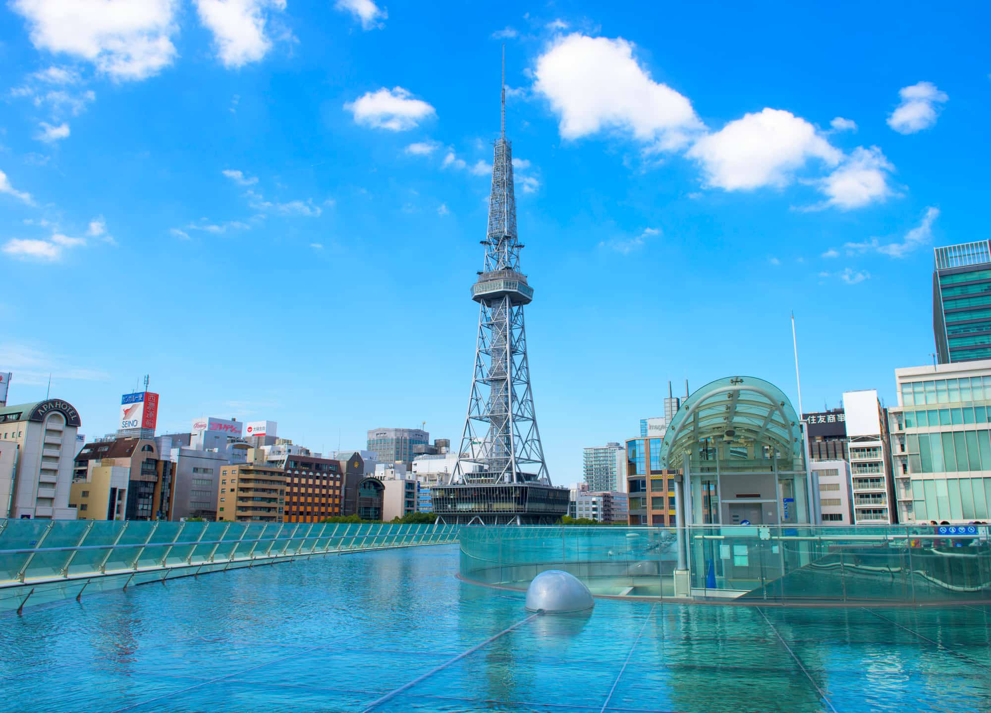Nagoya 桌布 壁紙 — 明亮的藍天白雲俯瞰著現代城市景觀，包括一座高鋼塔和一個玻璃屋頂建築，旁邊有一個反射池。
