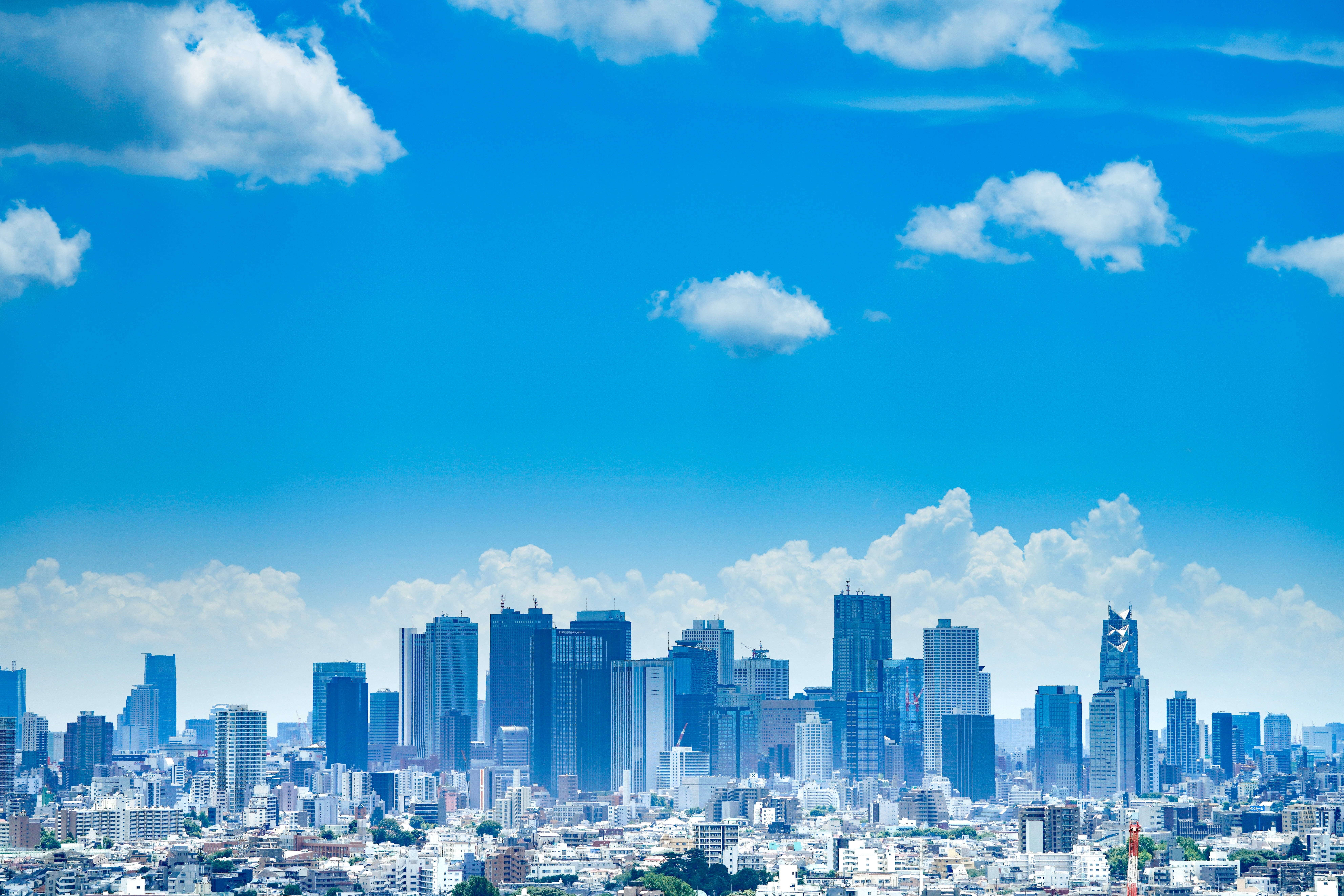 日本 桌布 壁紙 — 明亮的藍天與蓬鬆的白雲籠罩著密集的現代城市景觀，包括玻璃摩天大樓和低層建築。