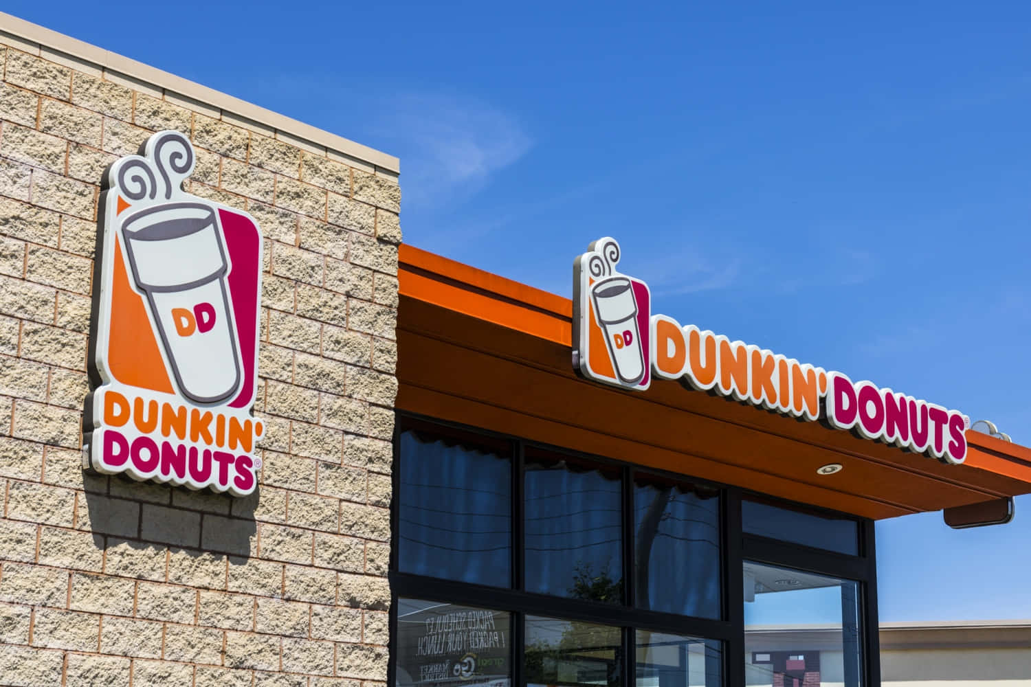 Dunkin Donuts 桌布 壁紙 — 明亮的藍天襯托著Dunkin' Donuts的店面，店面有醒目的橙色招牌和石磚牆。