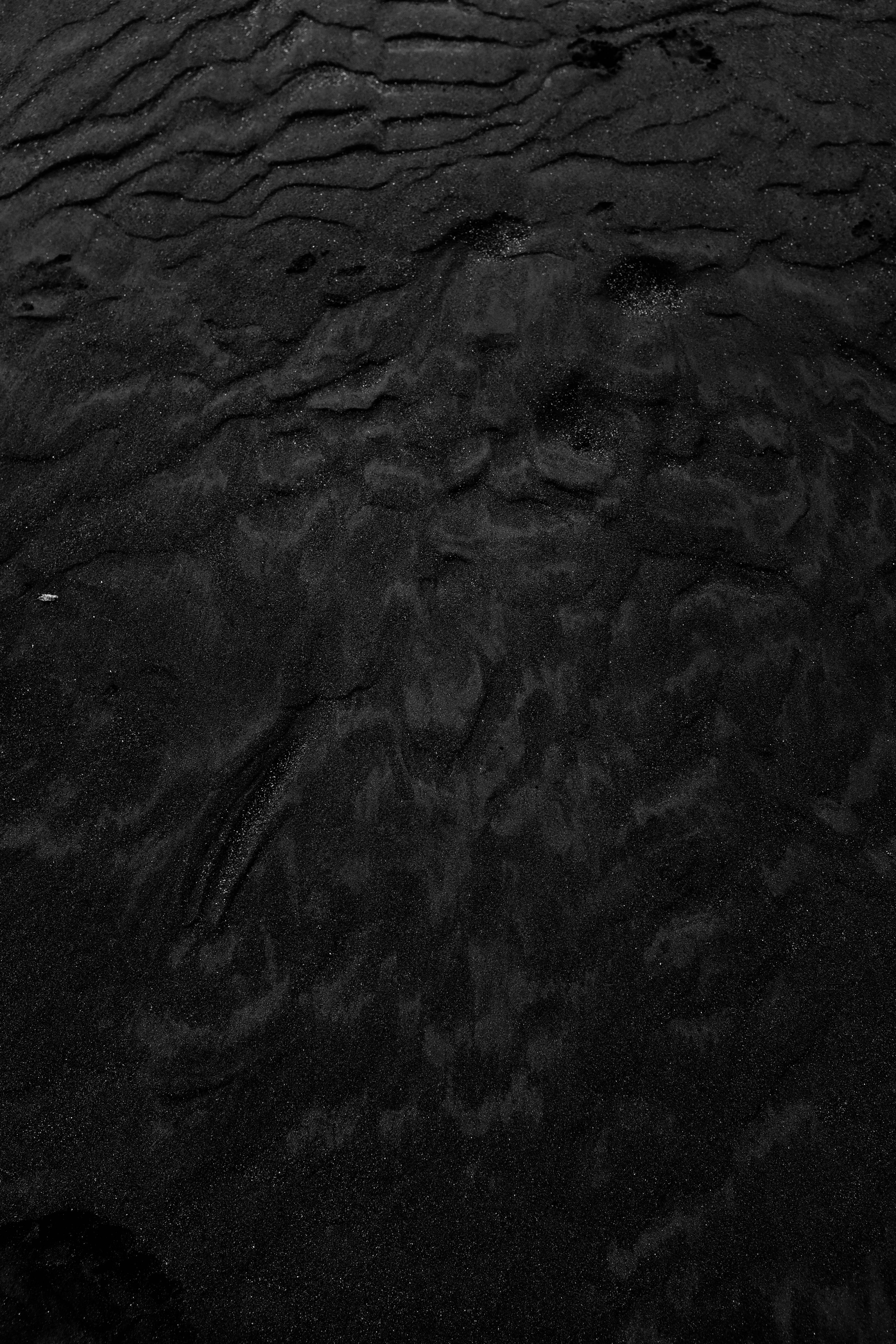 dark black 桌布 壁紙 — 昏暗光線下深色紋理沙子的特寫，帶有波浪狀漣漪和散落的小石頭。