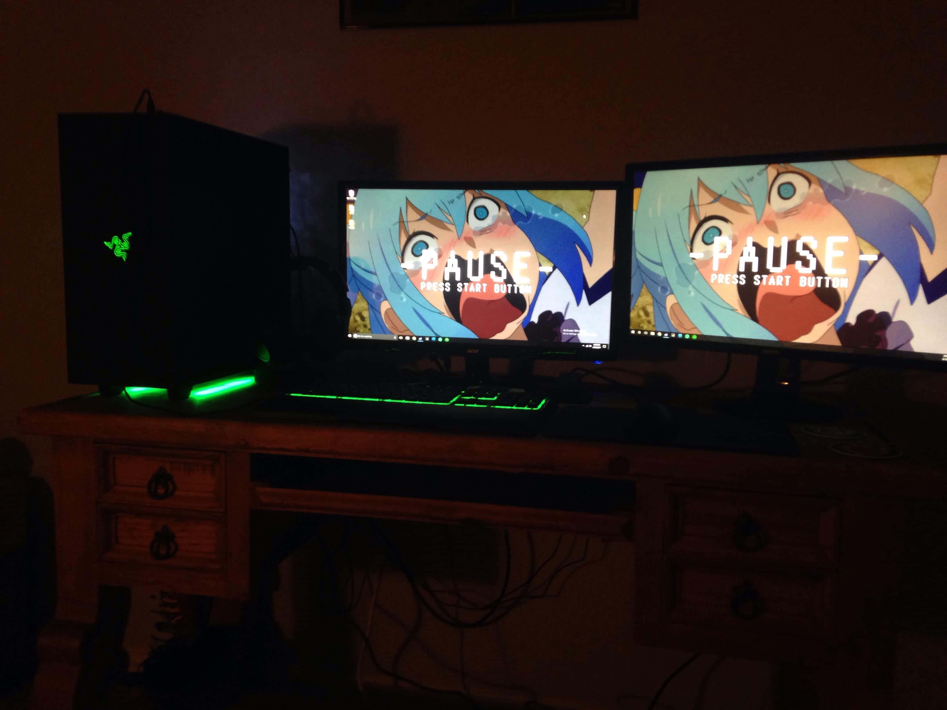 Gaming Setup 桌布 壁紙 — 昏暗燈光的遊戲設置，雙顯示器顯示動畫暫停畫面，木桌上有一個發光的綠色Razer PC塔。