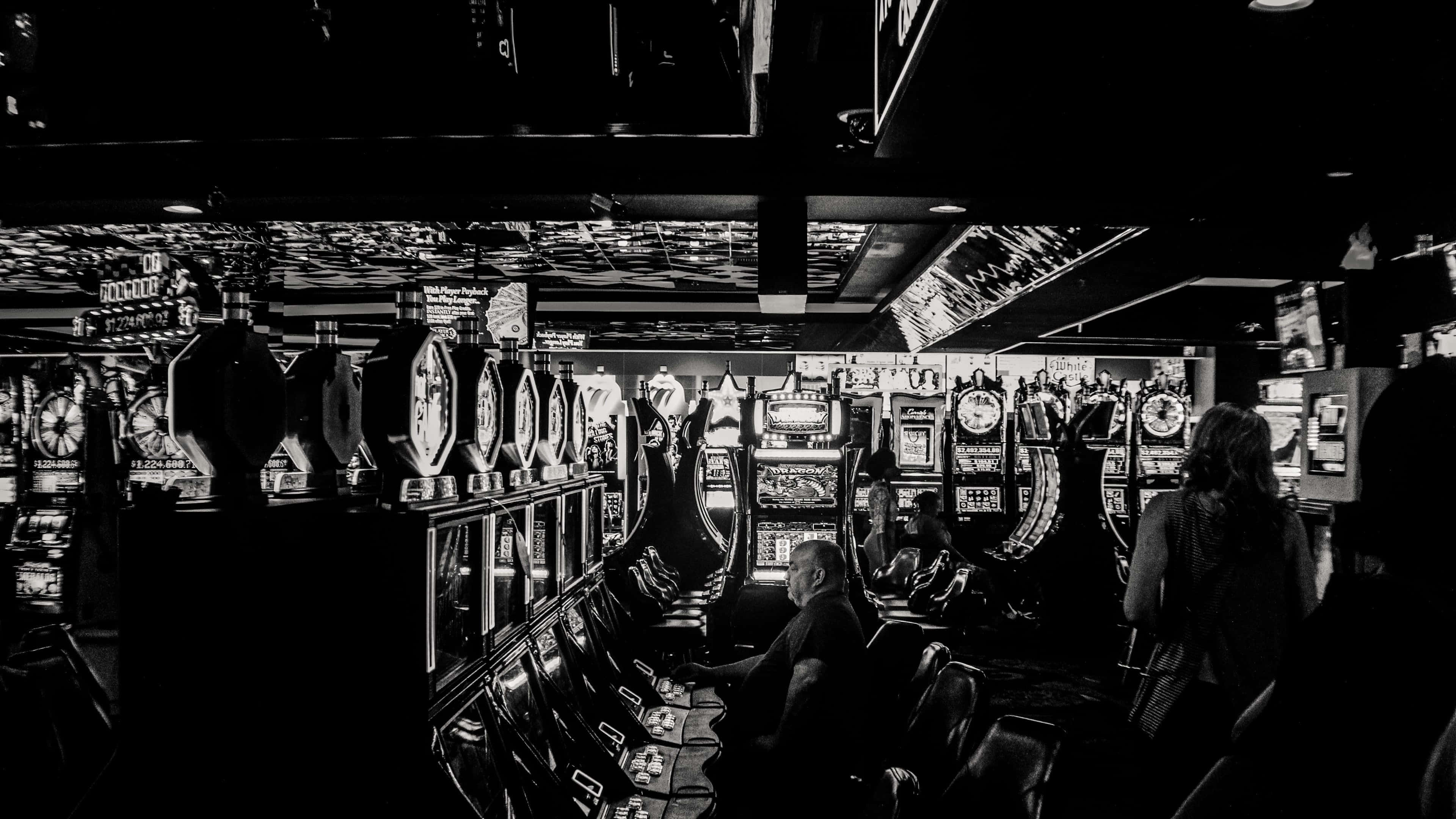 4K Las Vegas 桌布 壁紙 — 昏暗賭場中一排排的老虎機，顧客在發光的螢幕和反射天花板下玩樂。