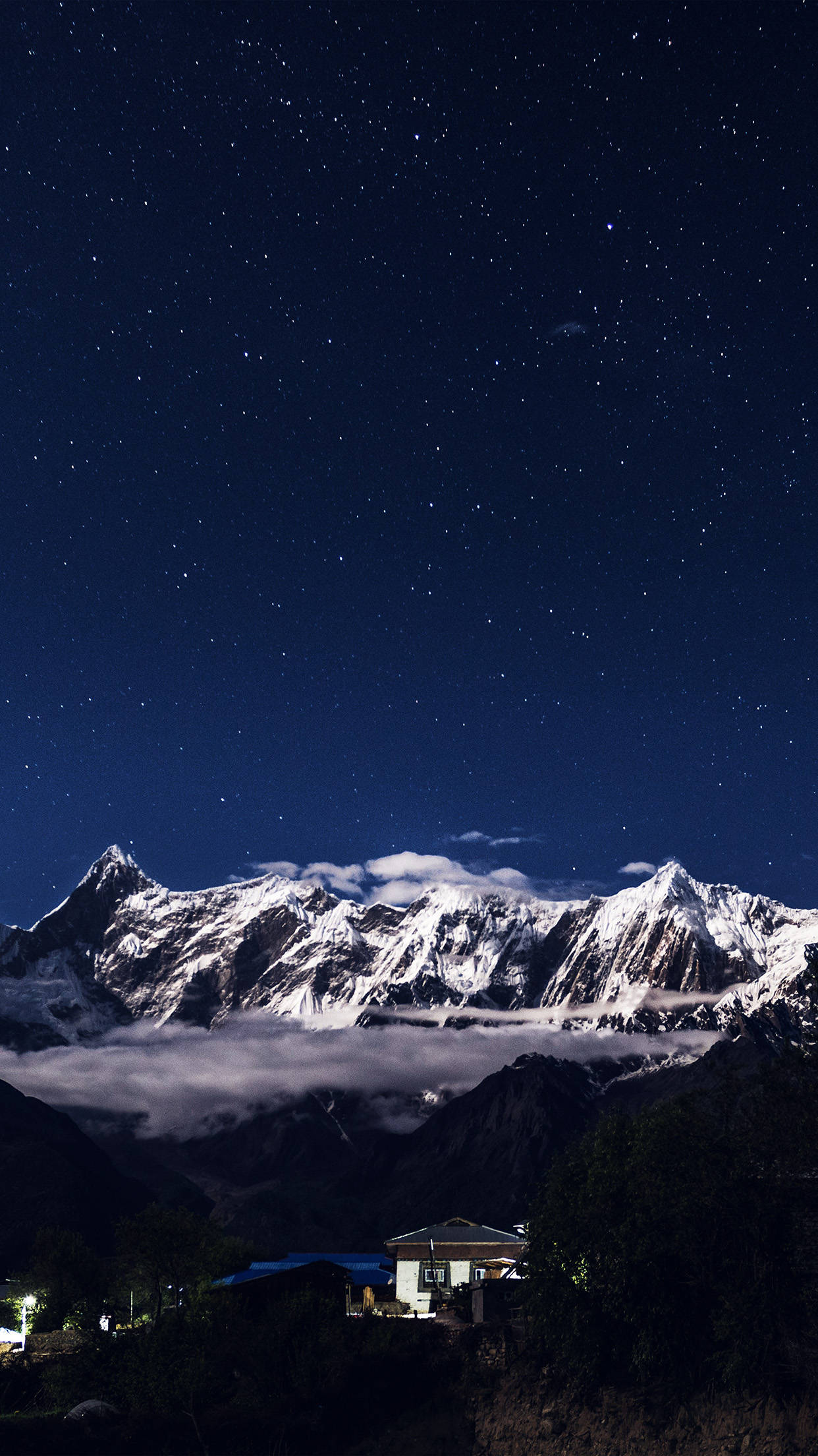 Night Mountain 桌布 壁紙 — 星空夜幕覆蓋著積雪的山峰，雲層在下，山谷中有一間亮燈的房子。