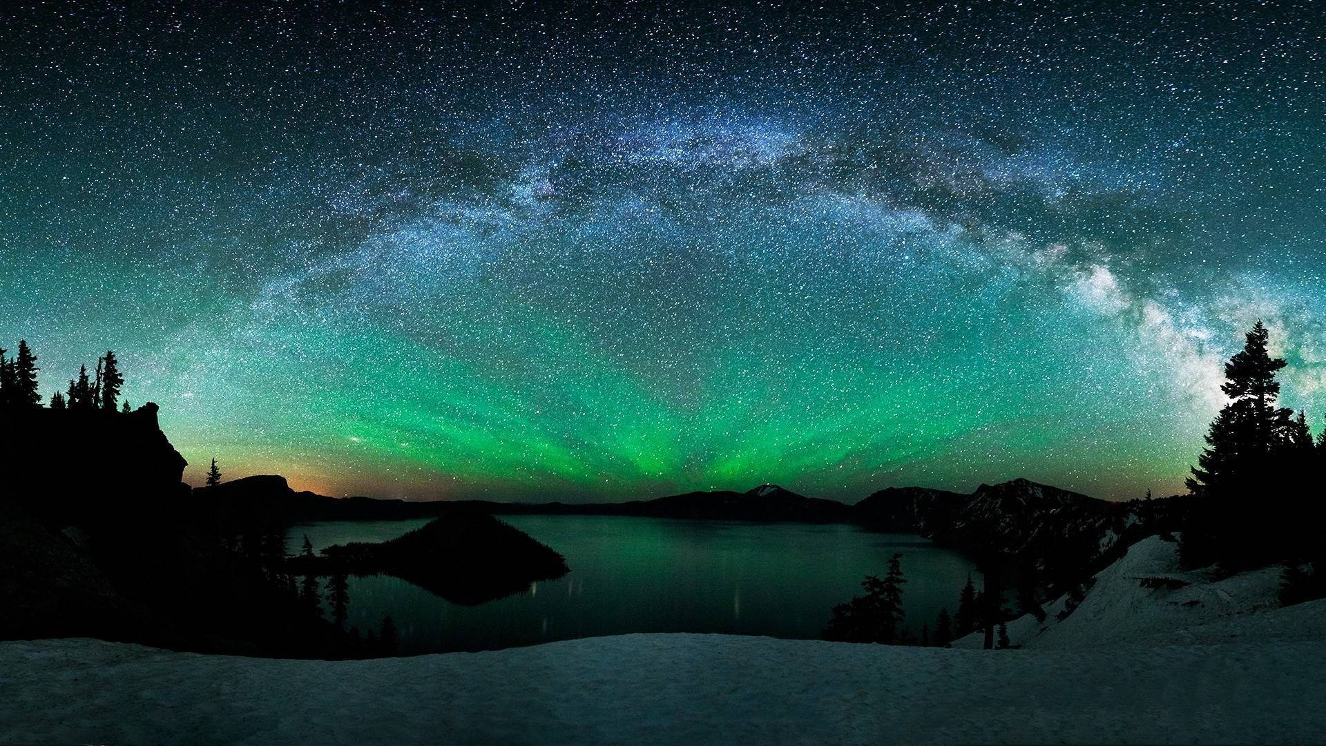 極光 桌布 壁紙 — 星空夜晚，銀河和綠色極光映照在白雪覆蓋的湖面上，周圍是黑暗的樹木。