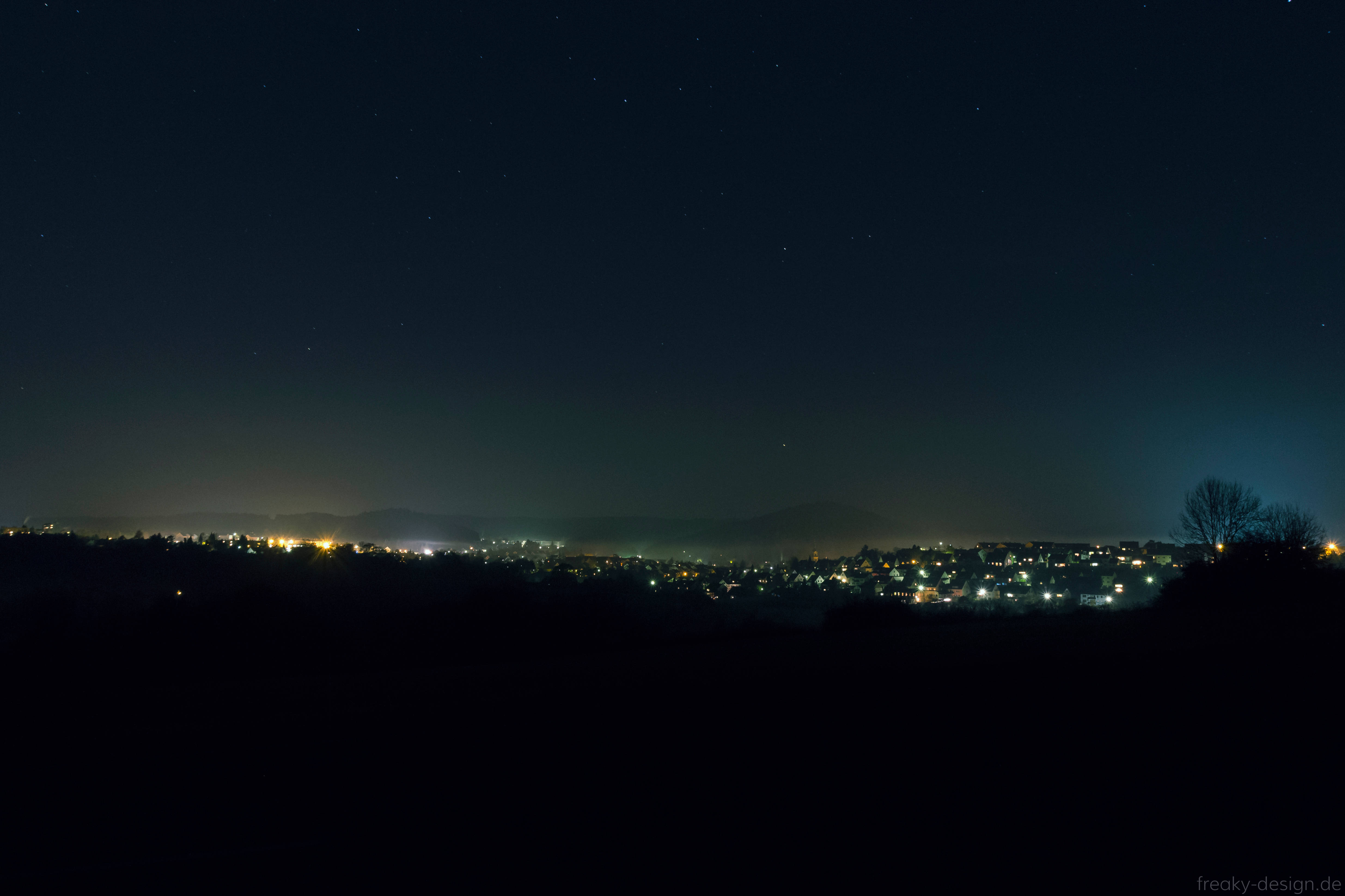 Starry 桌布 壁紙 — 星空夜空俯瞰著遠處的小鎮，在黑暗的山丘上散發著零星的黃色燈光。