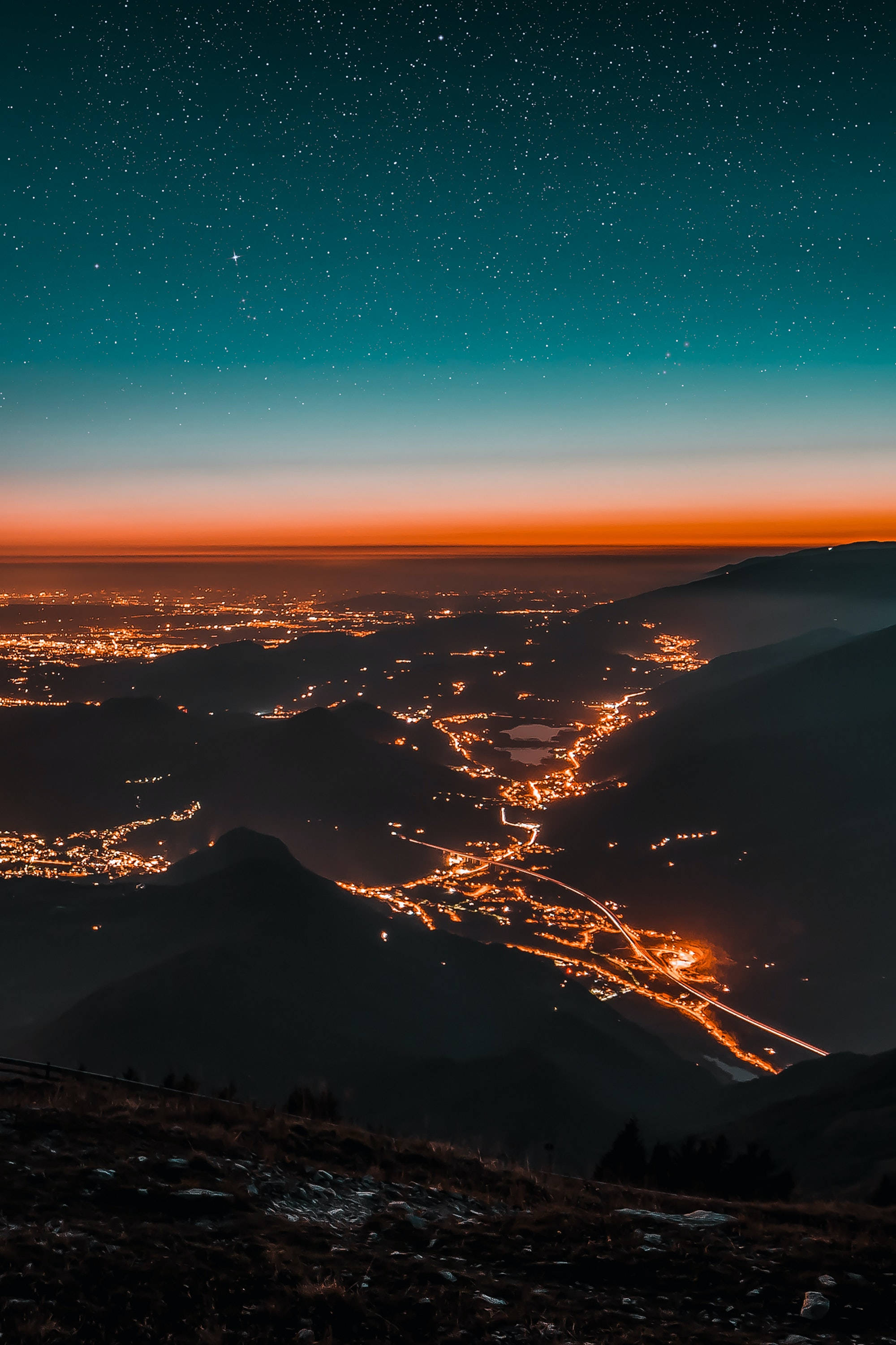 Light 桌布 壁紙 — 星空夜空在山谷小鎮上空發光，橙色燈光蜿蜒穿過黑暗的山脈。