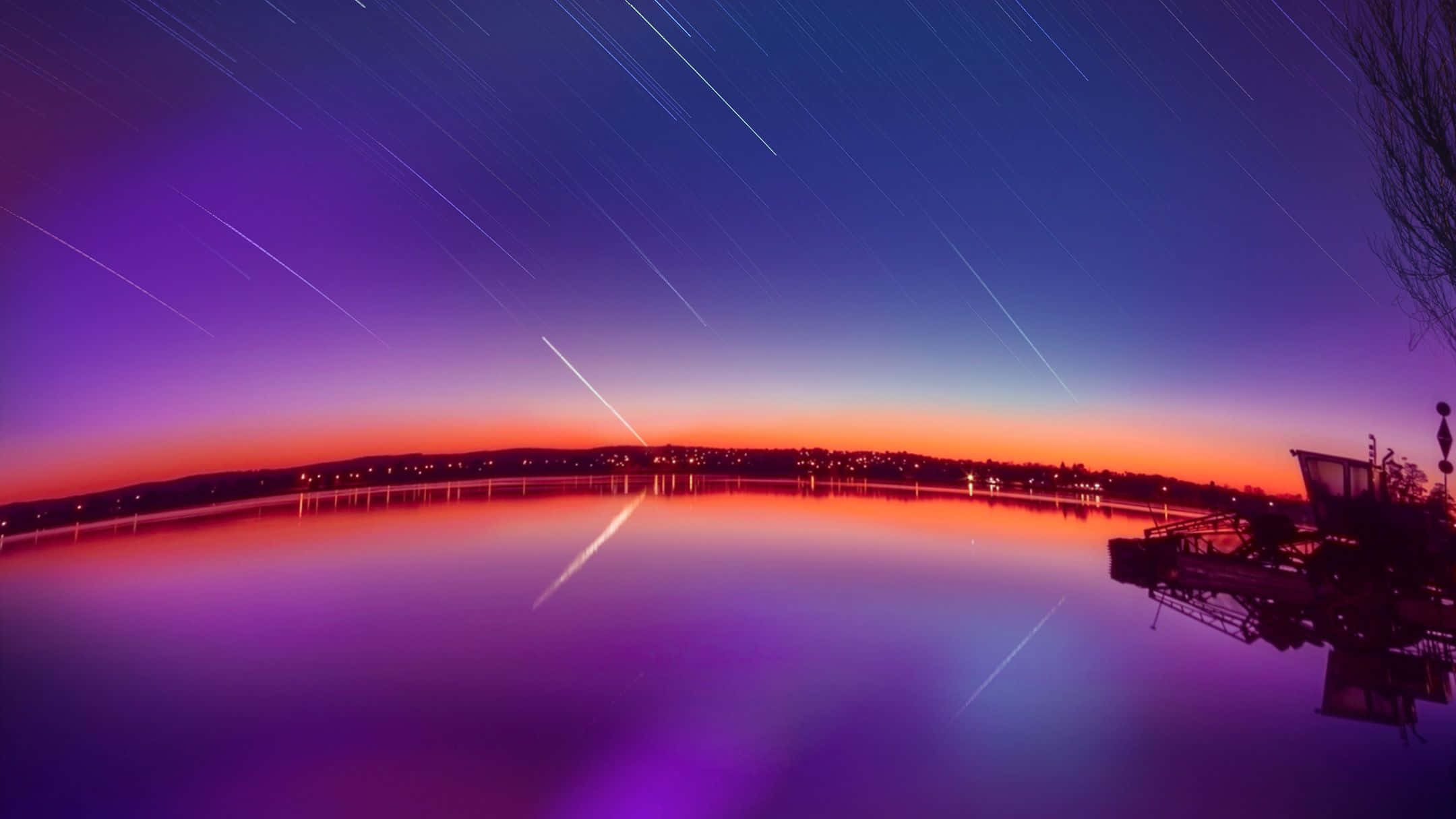Stars 桌布 壁紙 — 星軌劃過暮色天空，平靜的湖面反射出橙色和紫色的色調，遠處的城市燈光點綴其間。