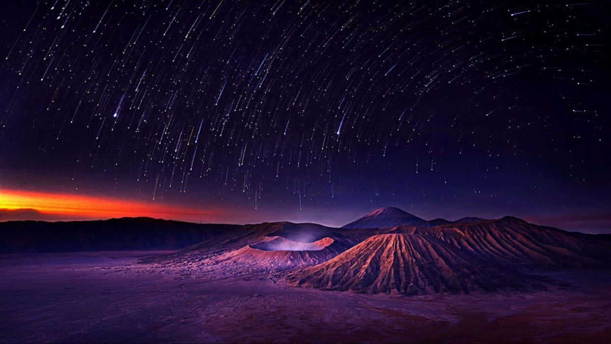 桌布 壁紙 — 星軌在深紫色和橙色光芒下，劃過崎嶇火山群山上空充滿活力的暮色天空。