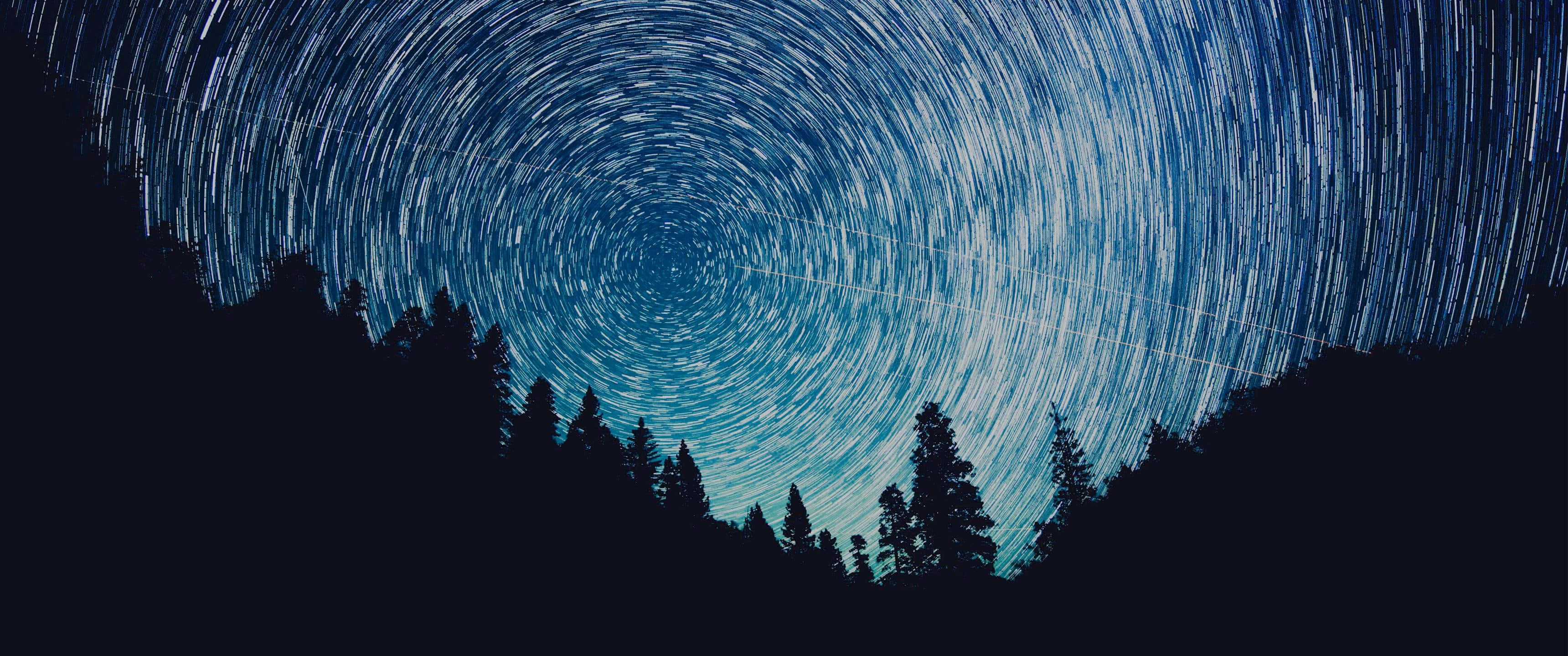 Ultra Wide 桌布 壁紙 — 星軌在深藍色夜空下，於黑暗森林剪影上方盤旋成迷人的弧線。