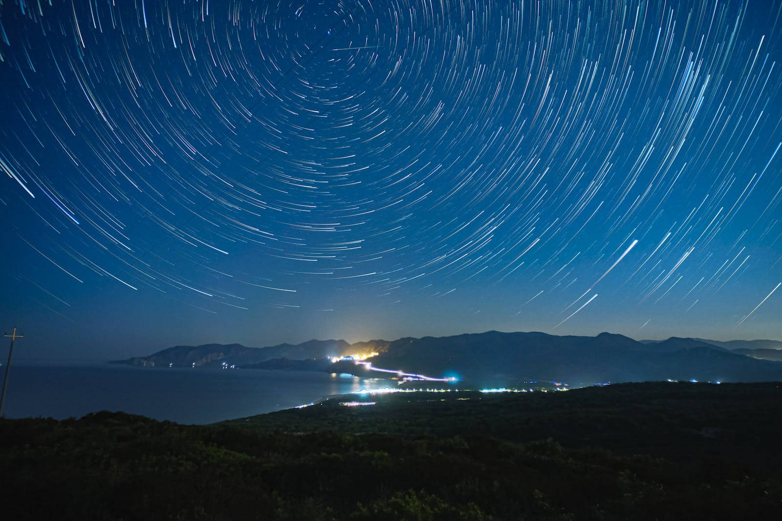 藍色銀河 桌布 壁紙 — 星軌在深藍色夜空中盤旋，下方是沿海小鎮，海岸線上有閃爍的燈光。