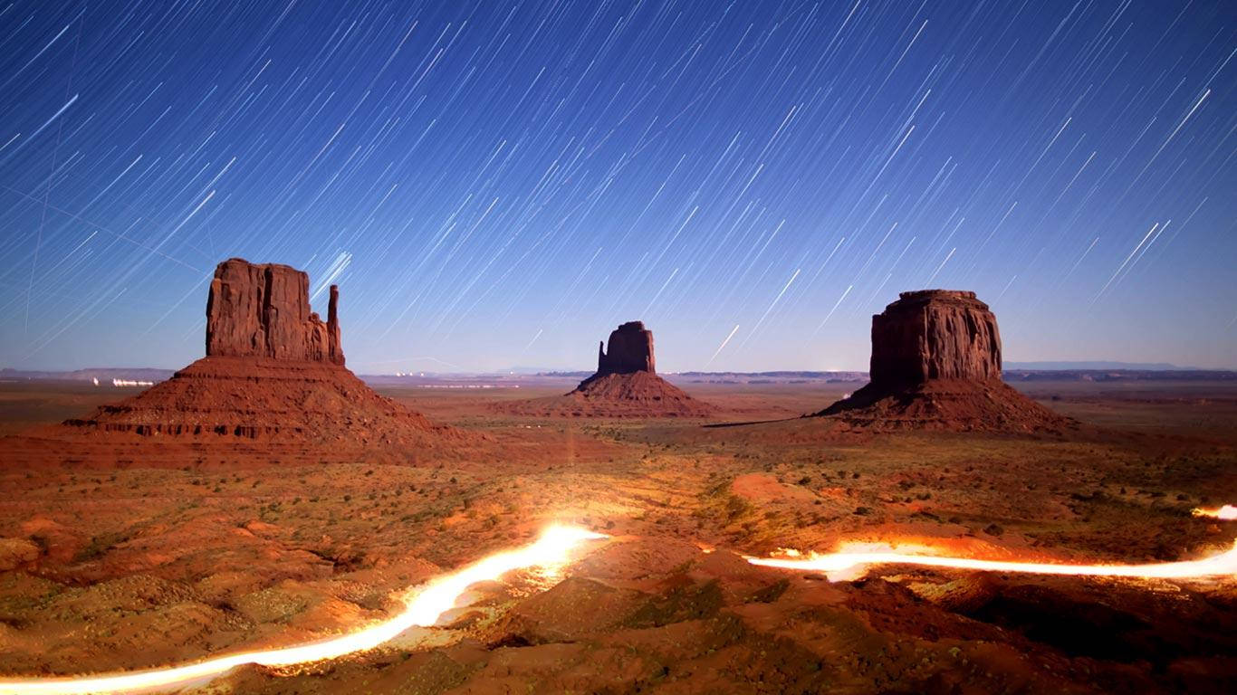 Stars 桌布 壁紙 — 星軌在深藍色的天空中劃過，映襯著標誌性的紅色岩層，發光的橙色光軌蜿蜒穿過沙漠地面。
