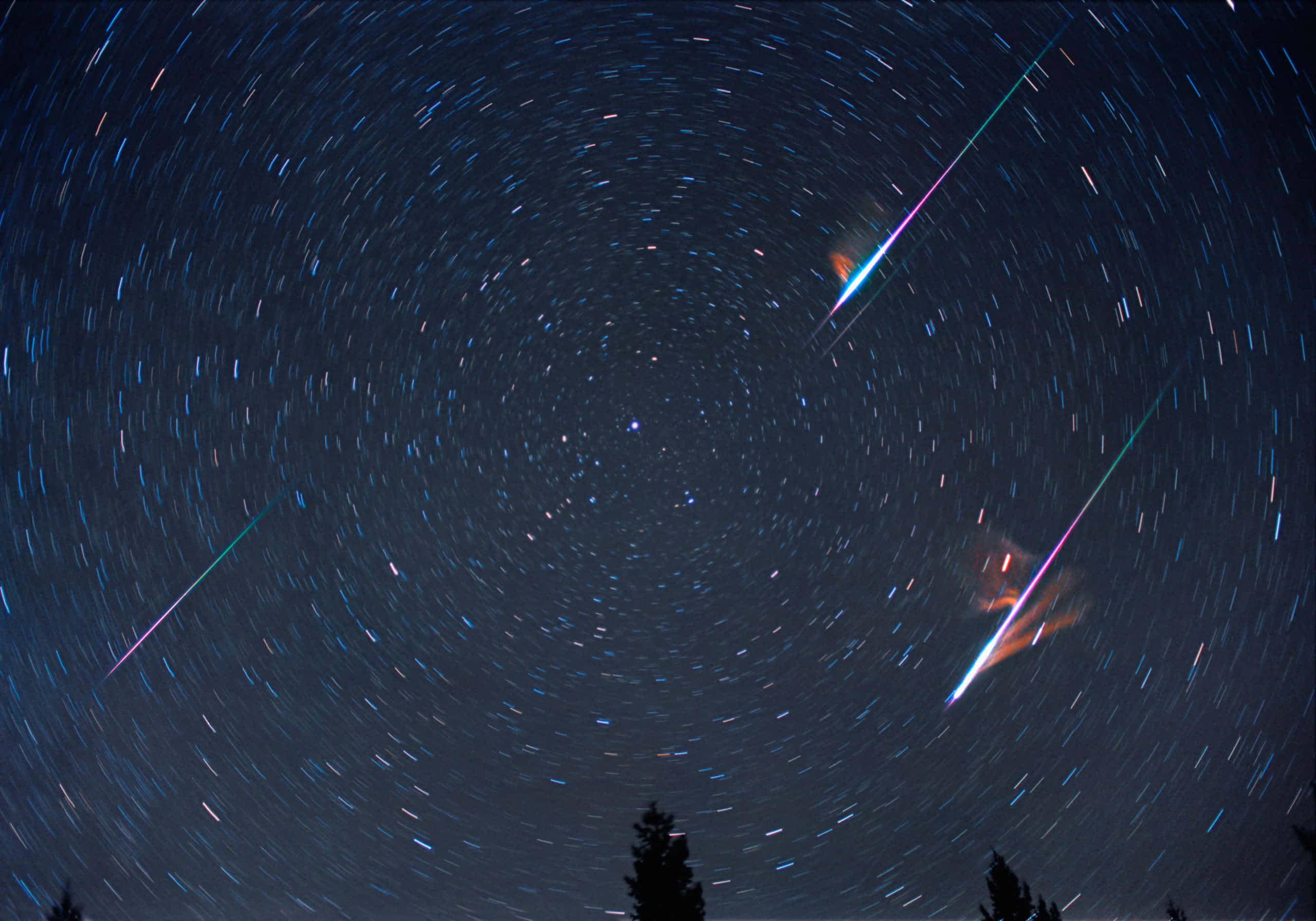 meteor 桌布 壁紙 — 星軌縮時攝影捕捉到兩顆鮮豔的流星劃過深藍色夜空，背景是剪影般的樹木。