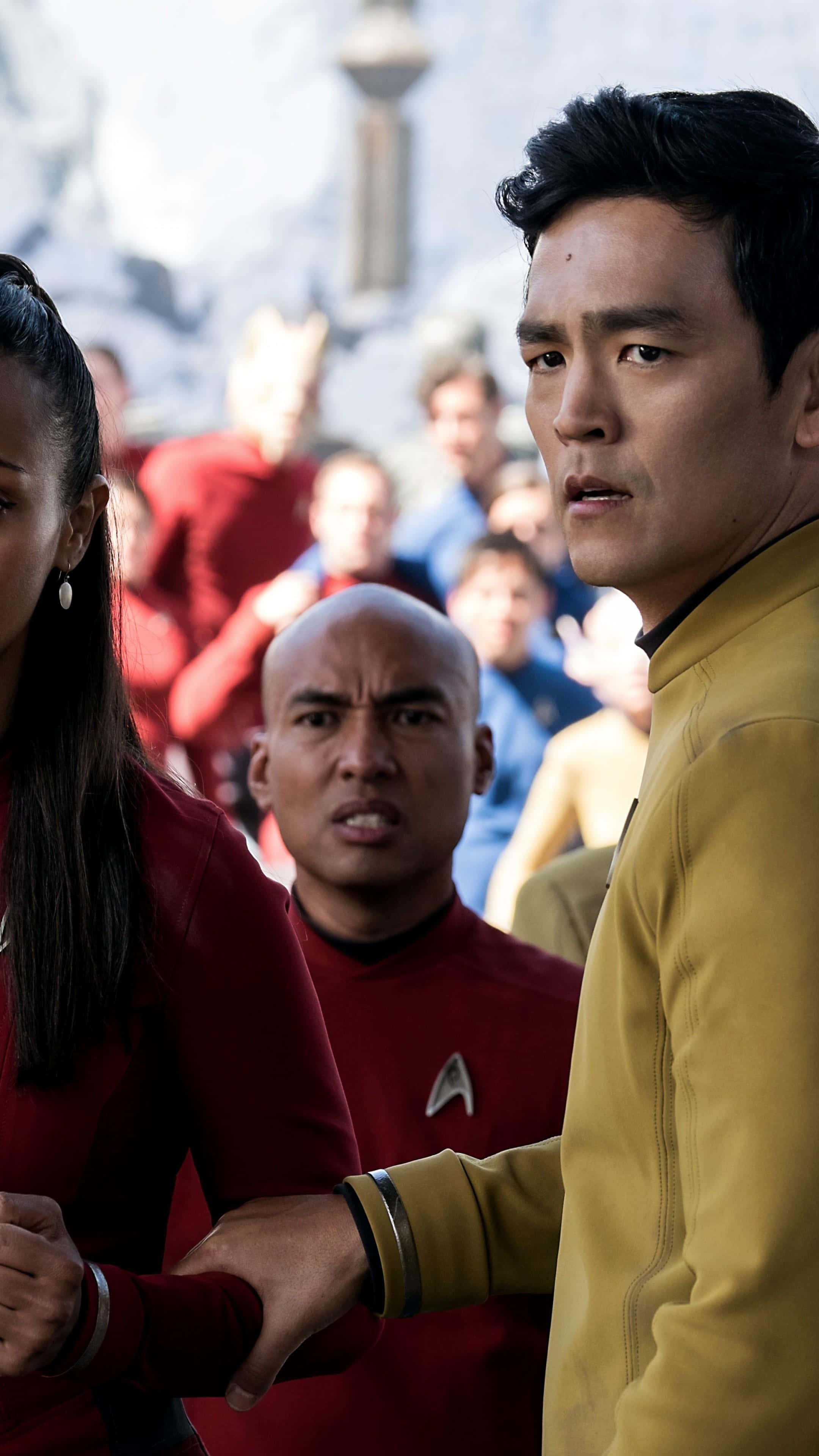John Cho 桌布 壁紙 — 《星際爭霸戰》船員穿著紅色和金色制服，在模糊的未來背景前緊張地站立。
