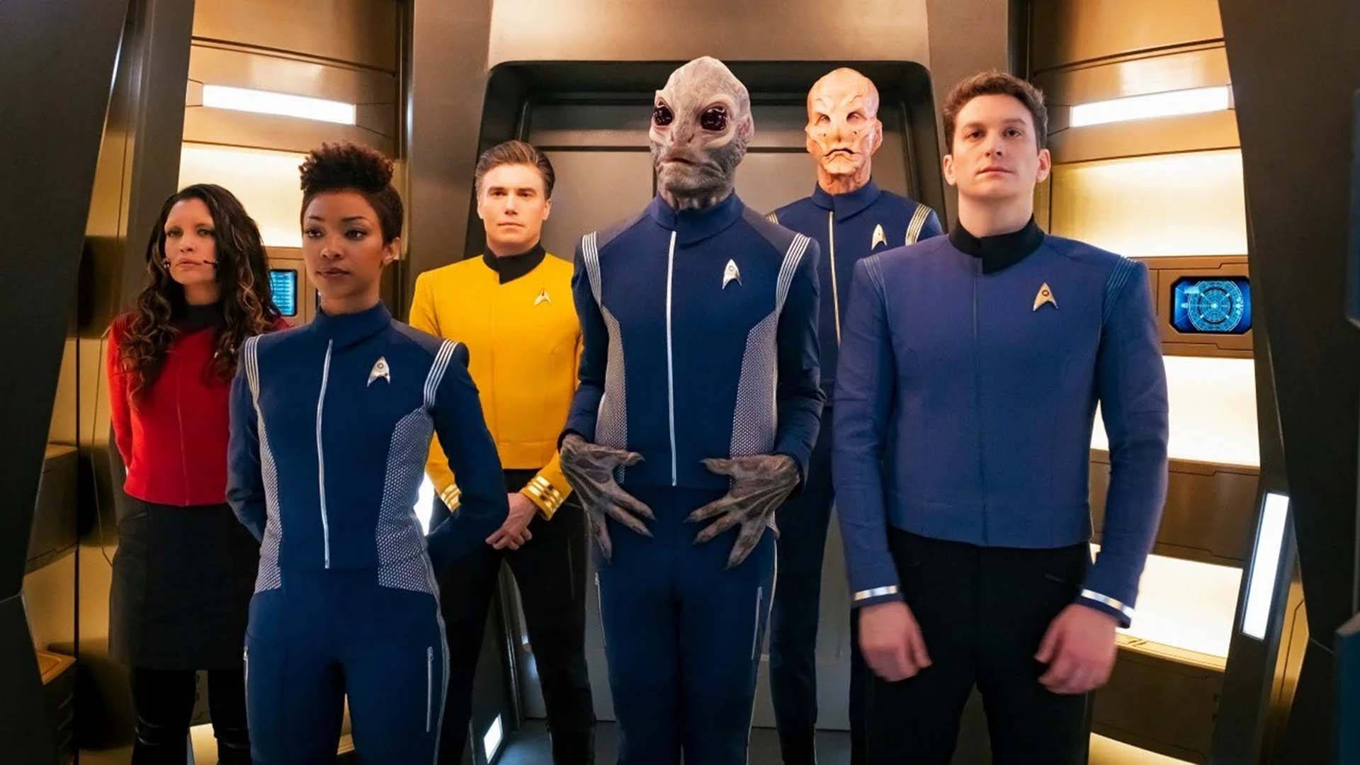 Discovery 桌布 壁紙 — 星際艦隊船員穿著色彩繽紛的制服，自信地站在未來主義的太空船走廊中，周圍有發光面板和外星船員。