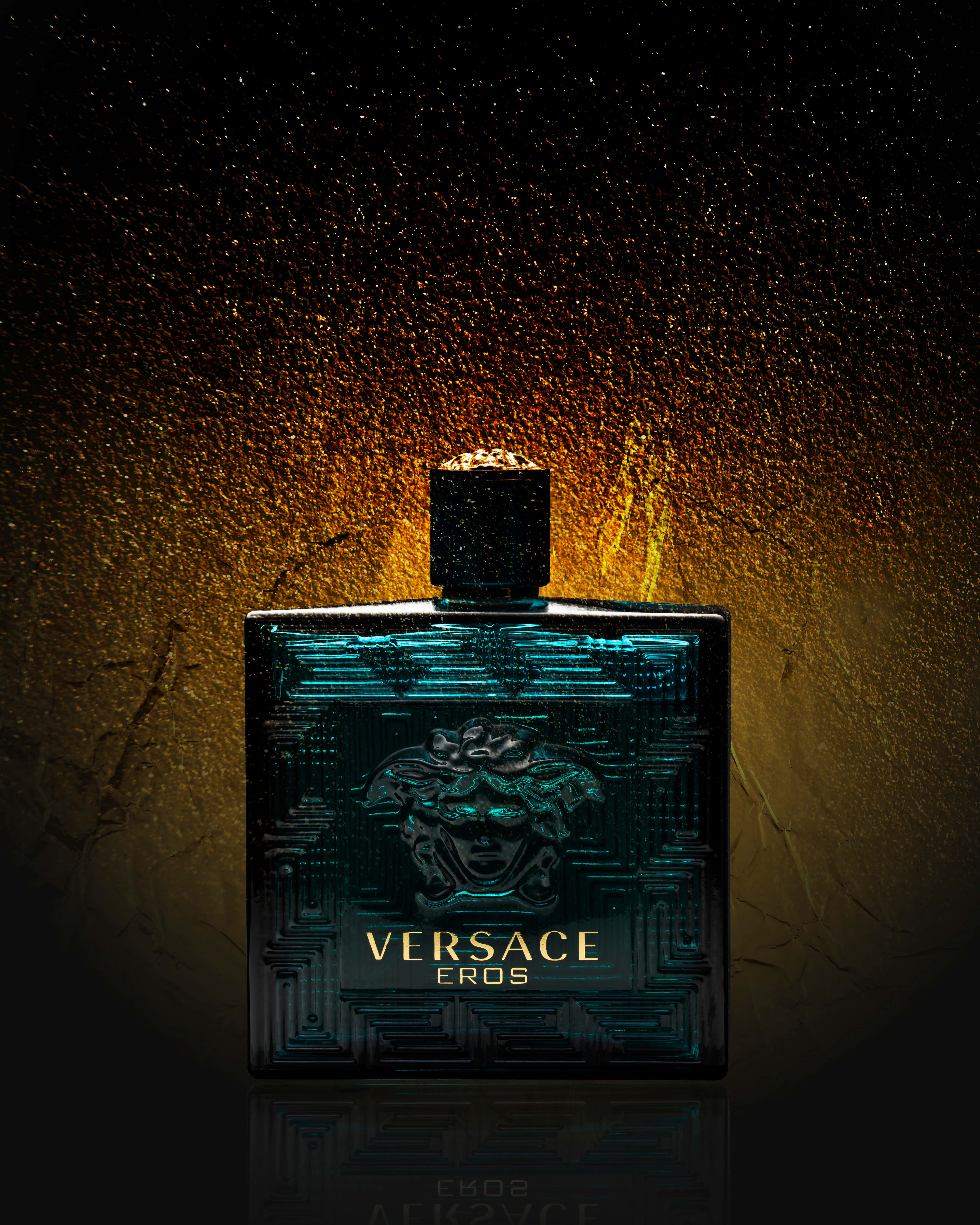 versace 桌布 壁紙 — 時尚的 Versace Eros 香水瓶在質感金色背景上閃爍著青綠色的光芒，背景上點綴著閃閃發光的顆粒。